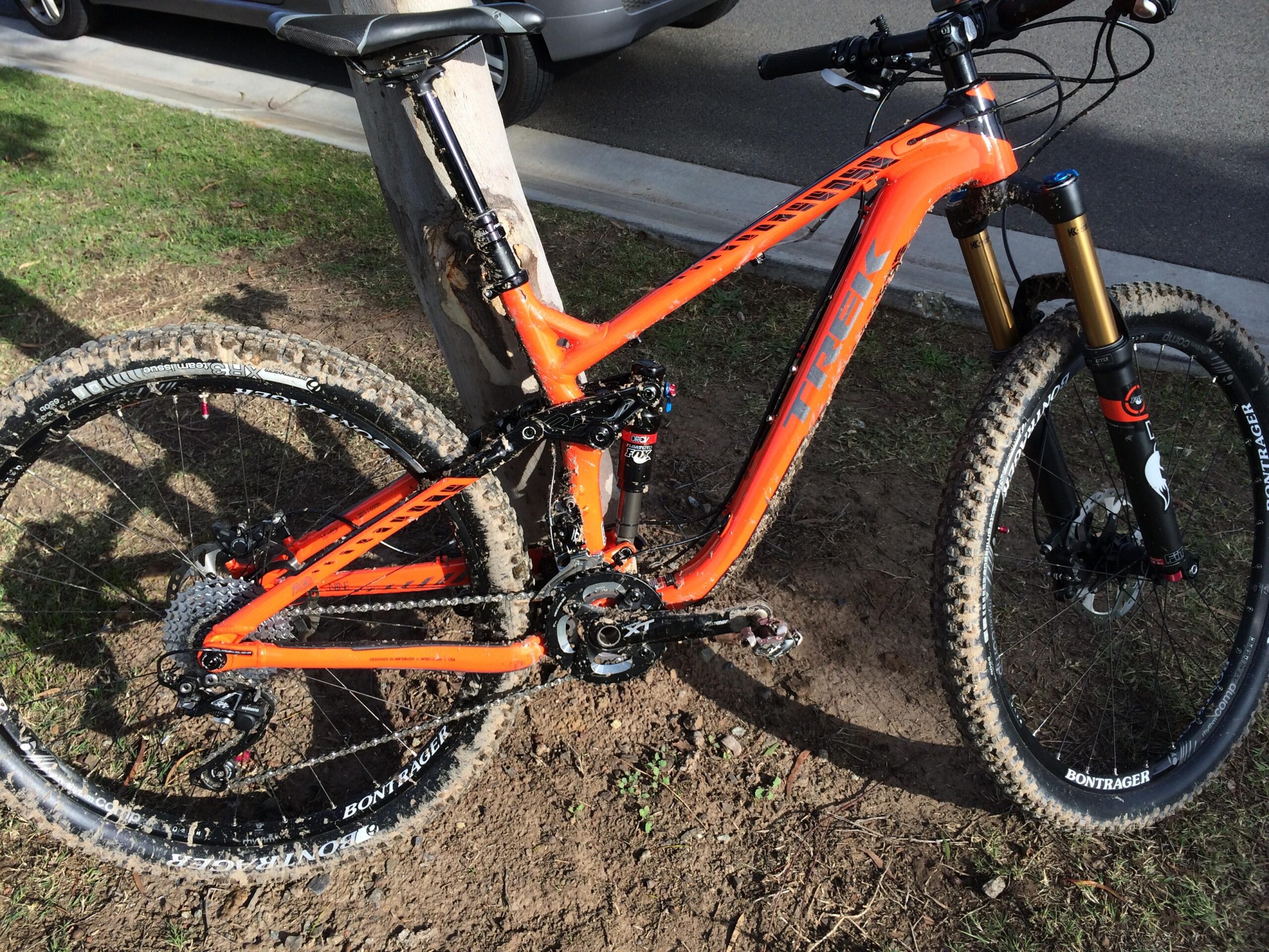 Trek Remedy 9: A close-up view of a bright orange mountain bike leaning against a wooden post. The bike features a modern design with thick tires, a dropper seat post, and a prominent brand logo. The frame is muddy, indicating recent outdoor use on trails or rugged terrain. The background shows a grassy area and part of a parked vehicle.
