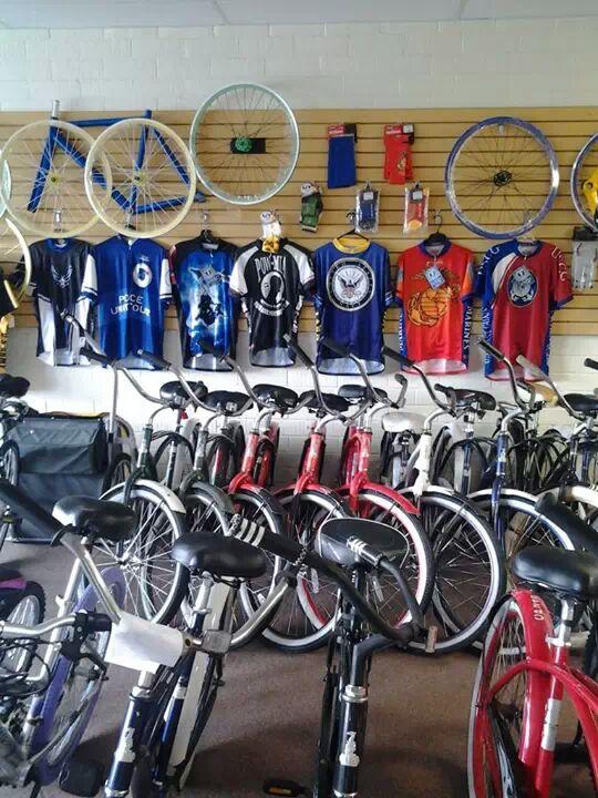 A display of various bicycles in a shop, with several bike jerseys hanging on the wall above. The bicycles include a mix of colors, primarily red and black, and are positioned closely together on the shop floor. The wall features bike wheels and various accessories, including socks and other biking gear.