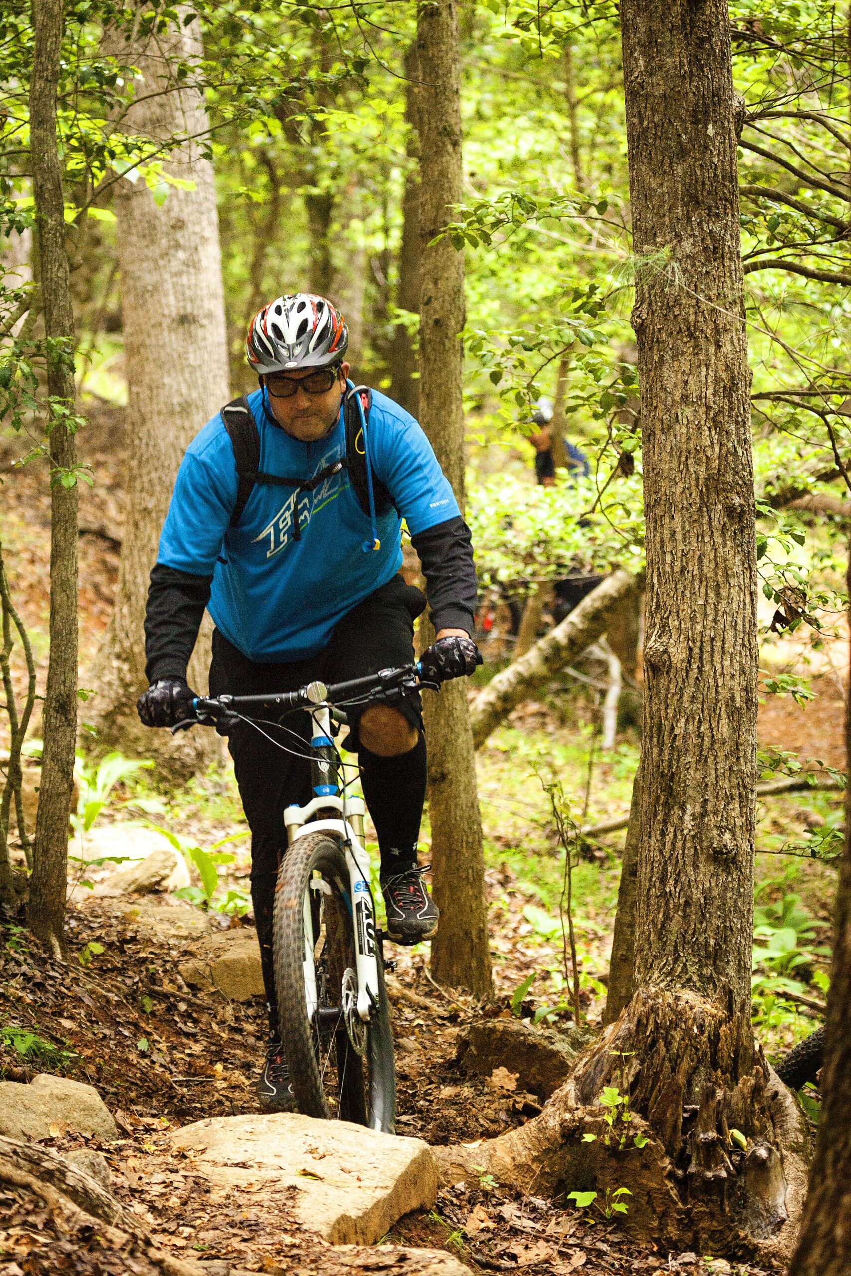 A mountain biker wearing a blue shirt, helmet, and gloves navigates a rocky trail surrounded by trees and greenery. Warrior Creek mountain bike trail.
