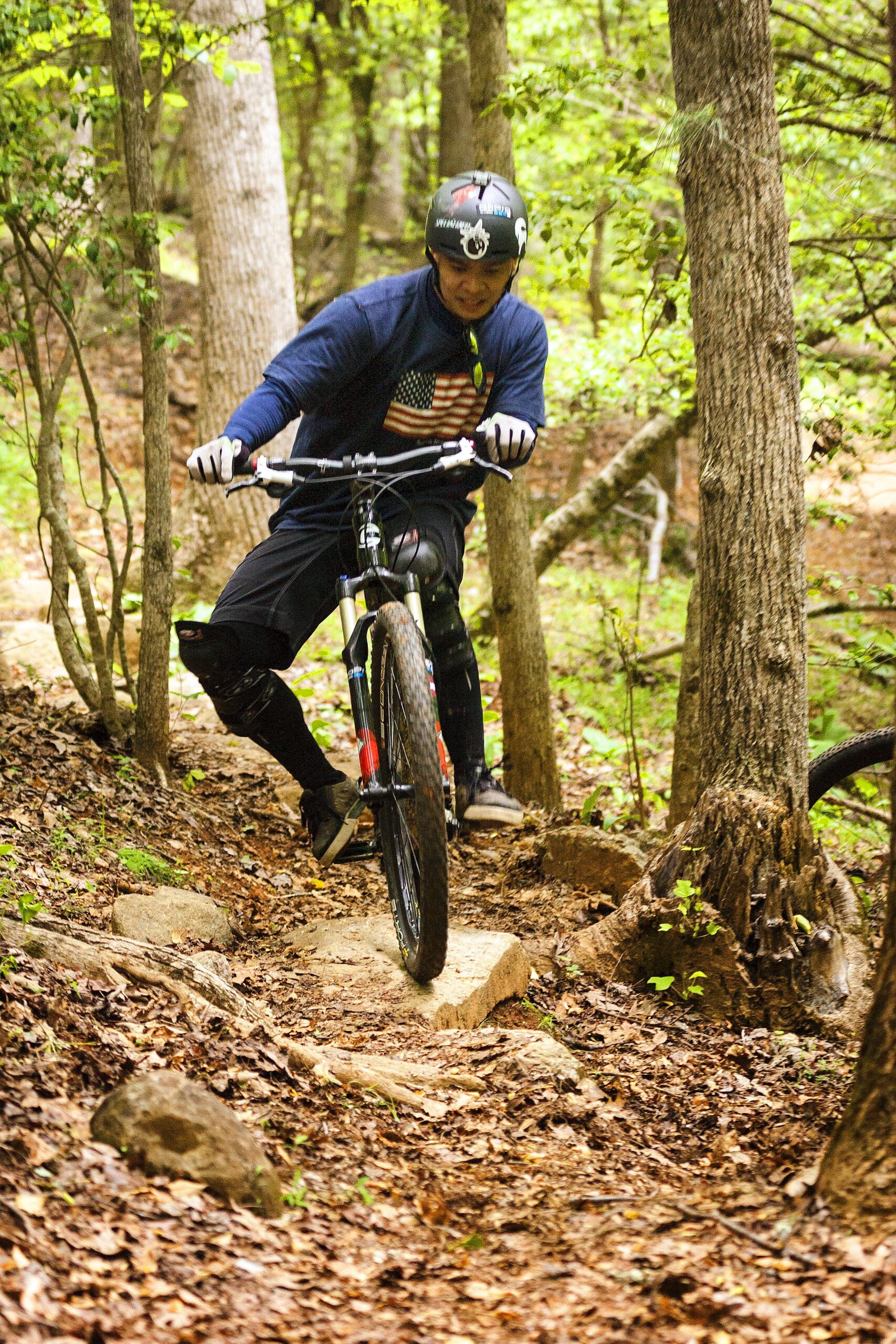 A mountain biker wearing a helmet and gloves rides over rocky terrain in a wooded area, surrounded by trees and fallen leaves. Warrior Creek mountain bike trail.