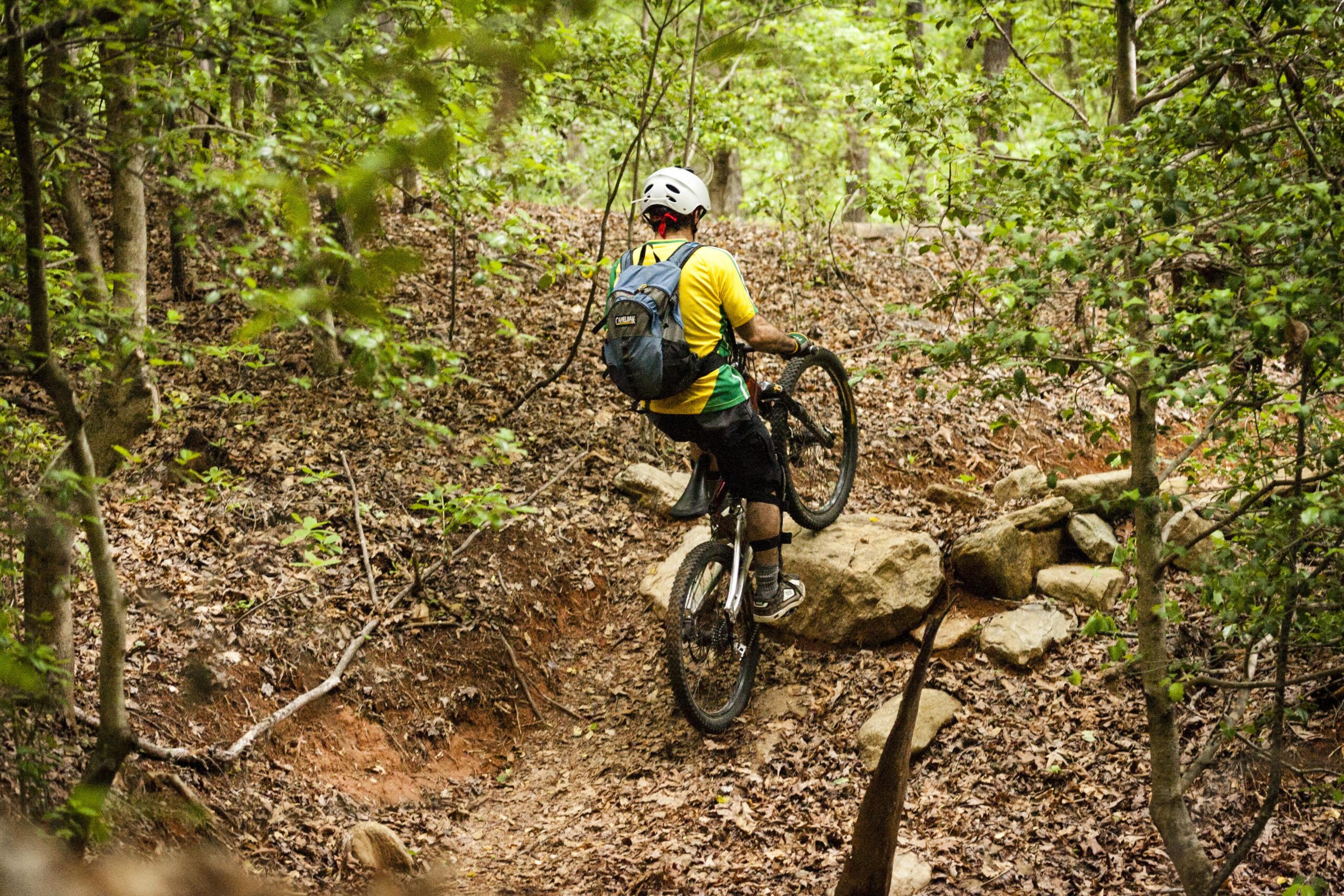 A mountain biker navigating a rocky trail in a wooded area, lifting the front wheel of the bike over a large stone, surrounded by green foliage and fallen leaves. The rider is wearing a helmet, a yellow and green shirt, and a backpack. Warrior Creek mountain bike trail.