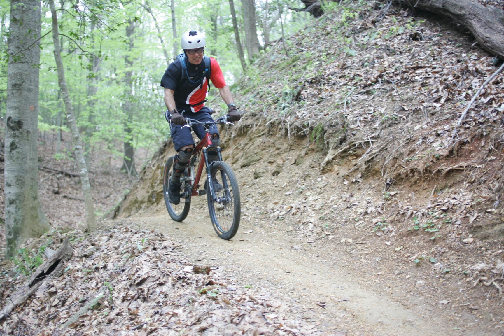 A mountain biker riding along a dirt trail in a lush green forest. The rider is wearing a helmet and protective gear, navigating the path surrounded by trees and fallen leaves. Warrior Creek mountain bike trail.