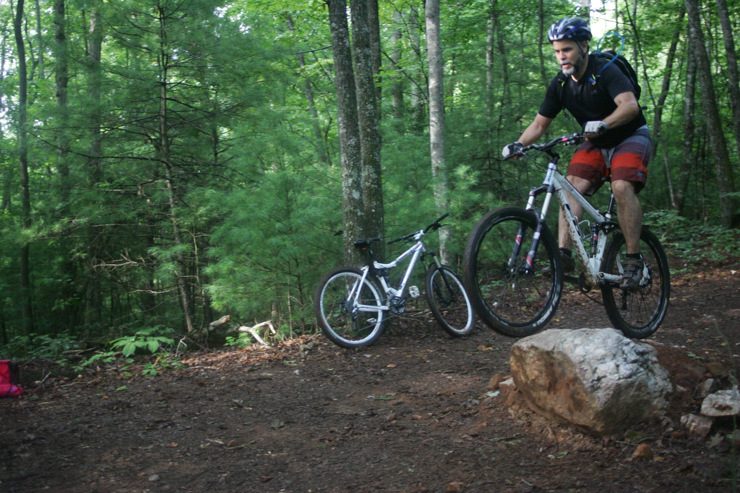 A mountain biker in colorful shorts and a helmet is mid-jump over a rock on a dirt trail, surrounded by dense green trees. A second bike is parked nearby. Dark Mountain Trail mountain bike trail.