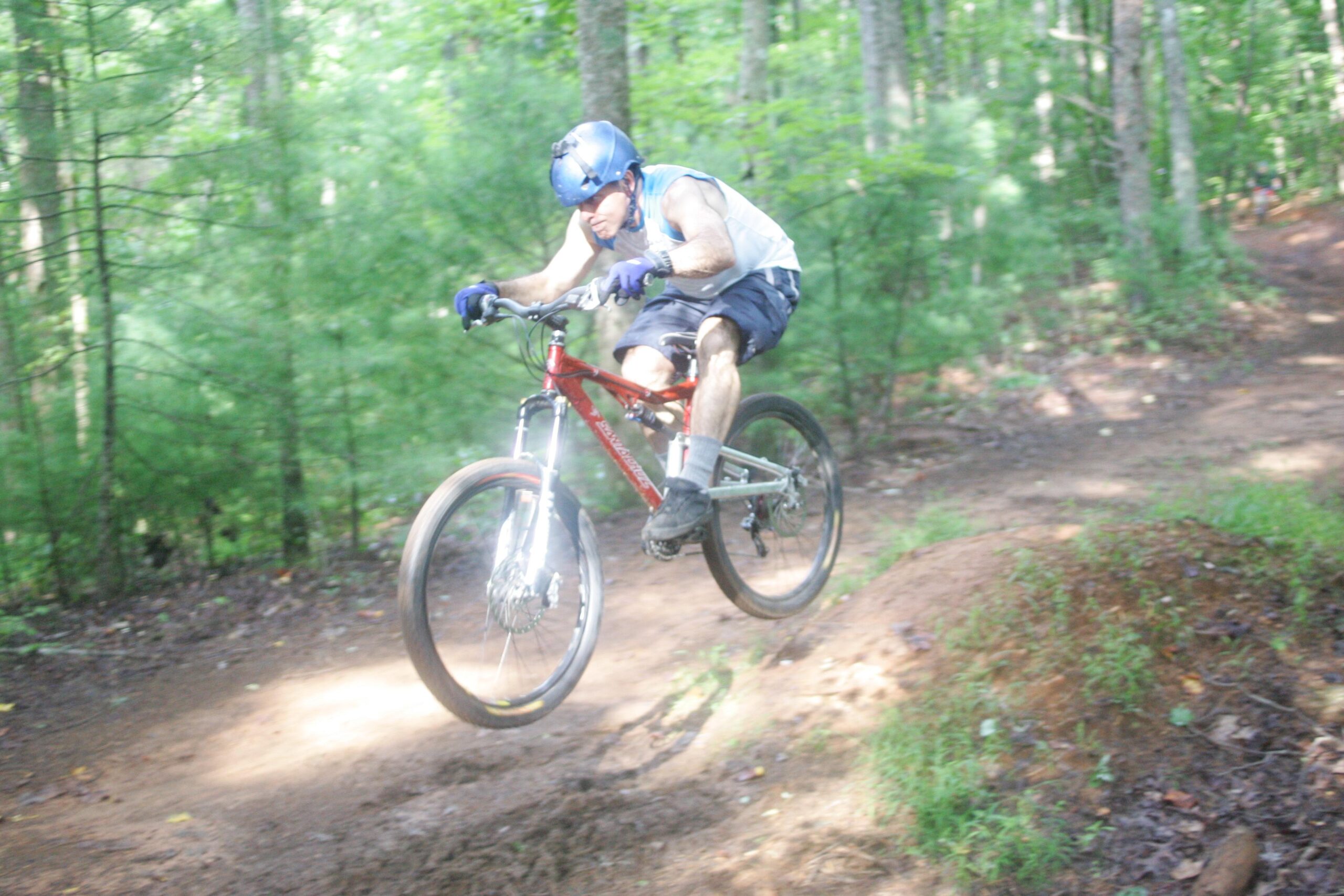A person in a helmet and gloves is riding a mountain bike on a dirt trail, mid-air after jumping off a small mound. The surrounding area is lush and green with trees, indicating a forested environment. The focus is on the cyclist’s dynamic action and the rugged terrain. Dark Mountain Trail mountain bike trail.
