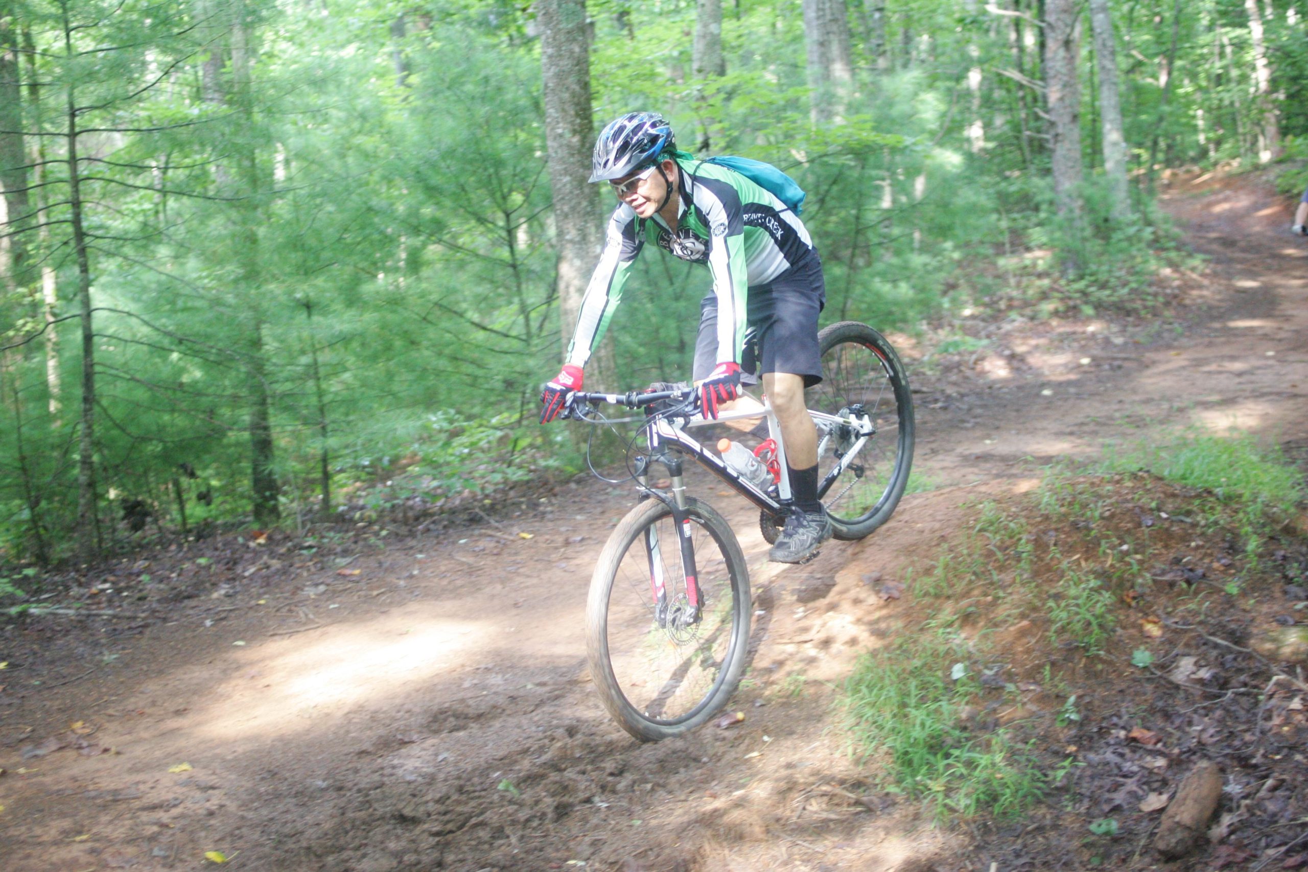 A mountain biker riding on a dirt trail surrounded by green trees, navigating a small incline with one wheel off the ground. The cyclist is wearing a helmet, gloves, and a sporty outfit, showcasing an action-packed moment in nature. Dark Mountain Trail mountain bike trail.