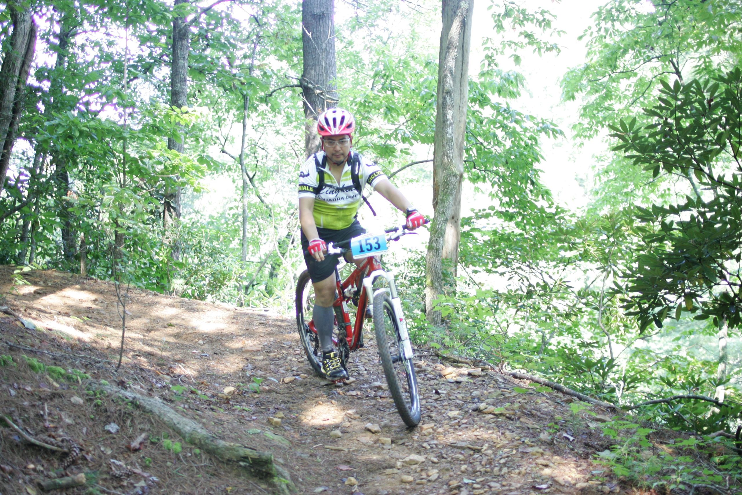 A mountain biker rides along a rocky trail surrounded by lush green trees. The cyclist, wearing a helmet and a brightly colored jersey with the number 153, is focused on navigating the terrain. Sunlight filters through the foliage, highlighting the vibrant natural surroundings. Dark Mountain Trail mountain bike trail.