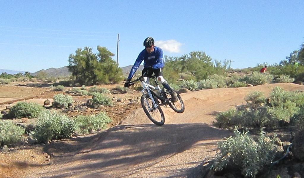 Giant Yukon FX: A mountain biker performs a jump on a dirt trail surrounded by desert vegetation under a clear blue sky.