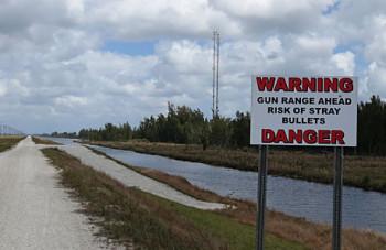 A gravel road leading to a gun range, with a warning sign that reads "WARNING GUN RANGE AHEAD RISK OF STRAY BULLETS DANGER." The background features a canal and trees under a cloudy sky. The Levee mountain bike trail.