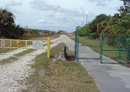 A gravel path leading to a green gate, flanked by yellow and white warning signs. Lush greenery is visible on either side, under a partly cloudy sky. The Levee mountain bike trail.