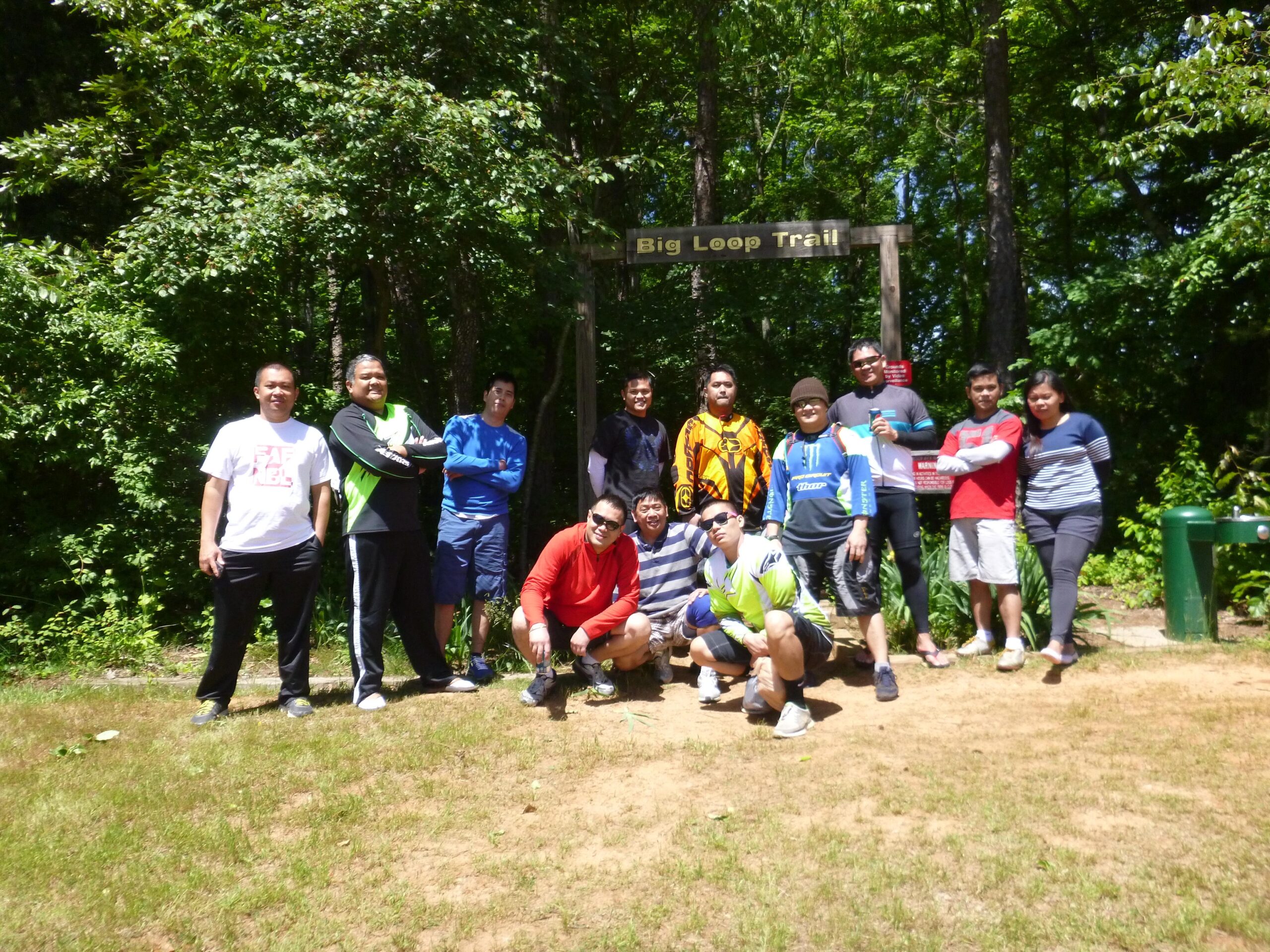 Group of individuals gathered at the Big Loop Trail sign in a wooded area, smiling and posing for the photo. They are wearing casual outdoor clothing, and the scene is sunny with lush greenery in the background. Big Loop mountain bike trail.