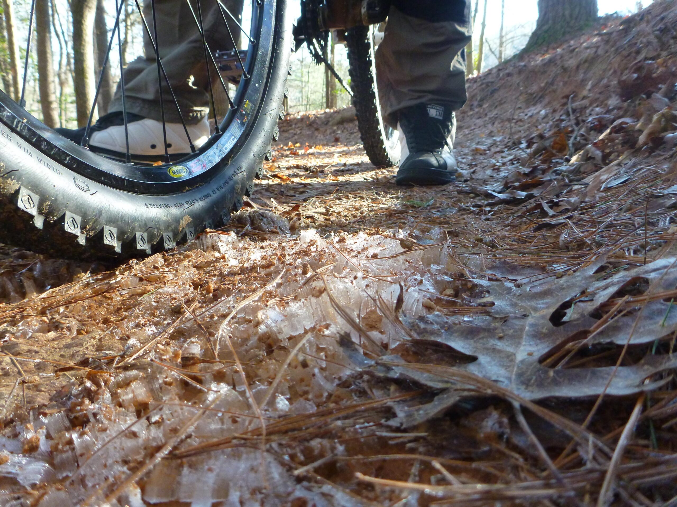 Close-up of a mountain bike tire on a muddy trail covered with fallen leaves and patches of ice. A person's leg is visible in the background, wearing sturdy footwear. Sunlight filters through the trees, highlighting the natural setting. Overmountain Victory Trail mountain bike trail.