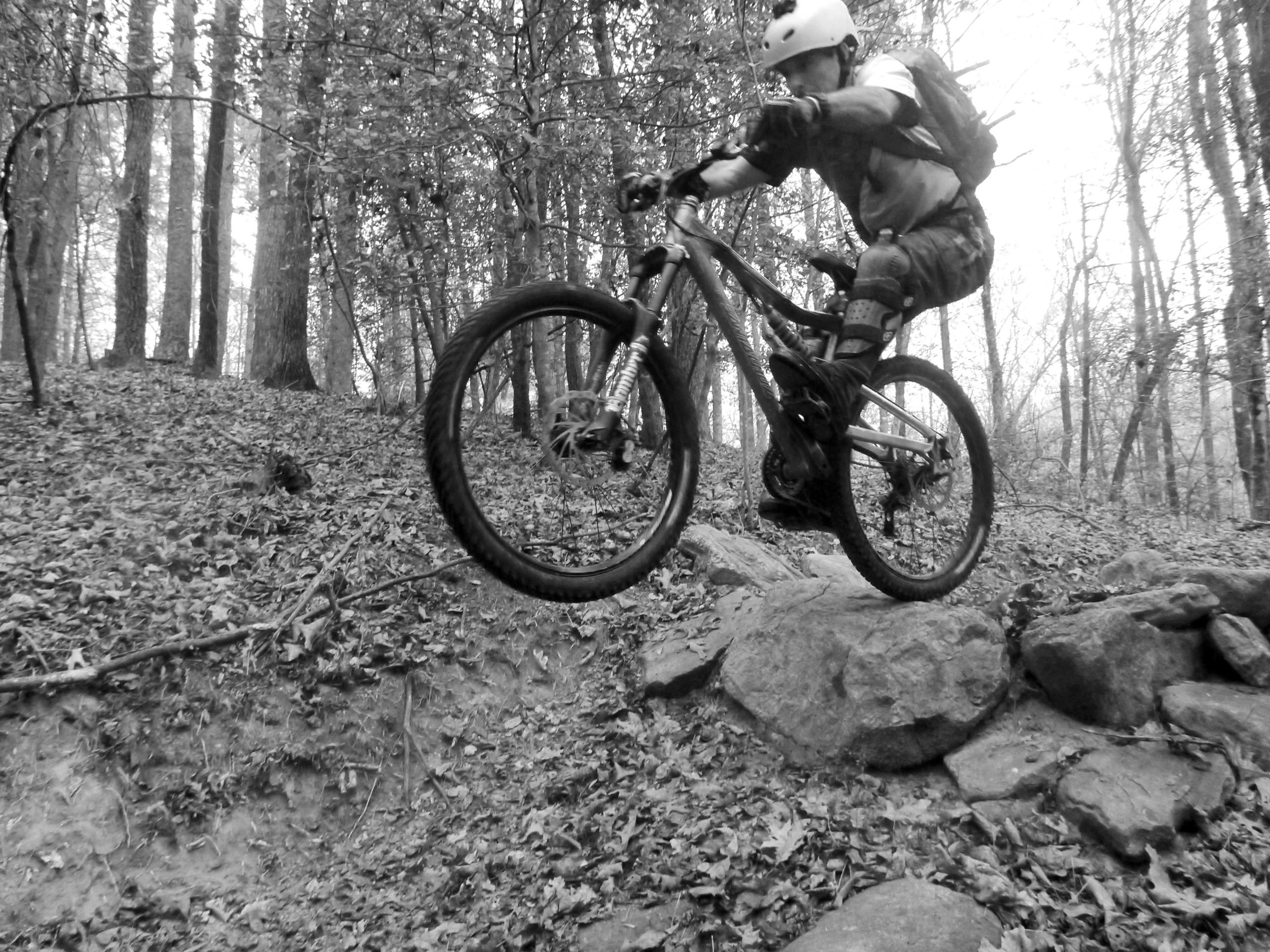A mountain biker in a helmet and protective gear jumps off a rocky ledge in a forest, surrounded by trees and fallen leaves, captured in black and white. Warrior Creek mountain bike trail.