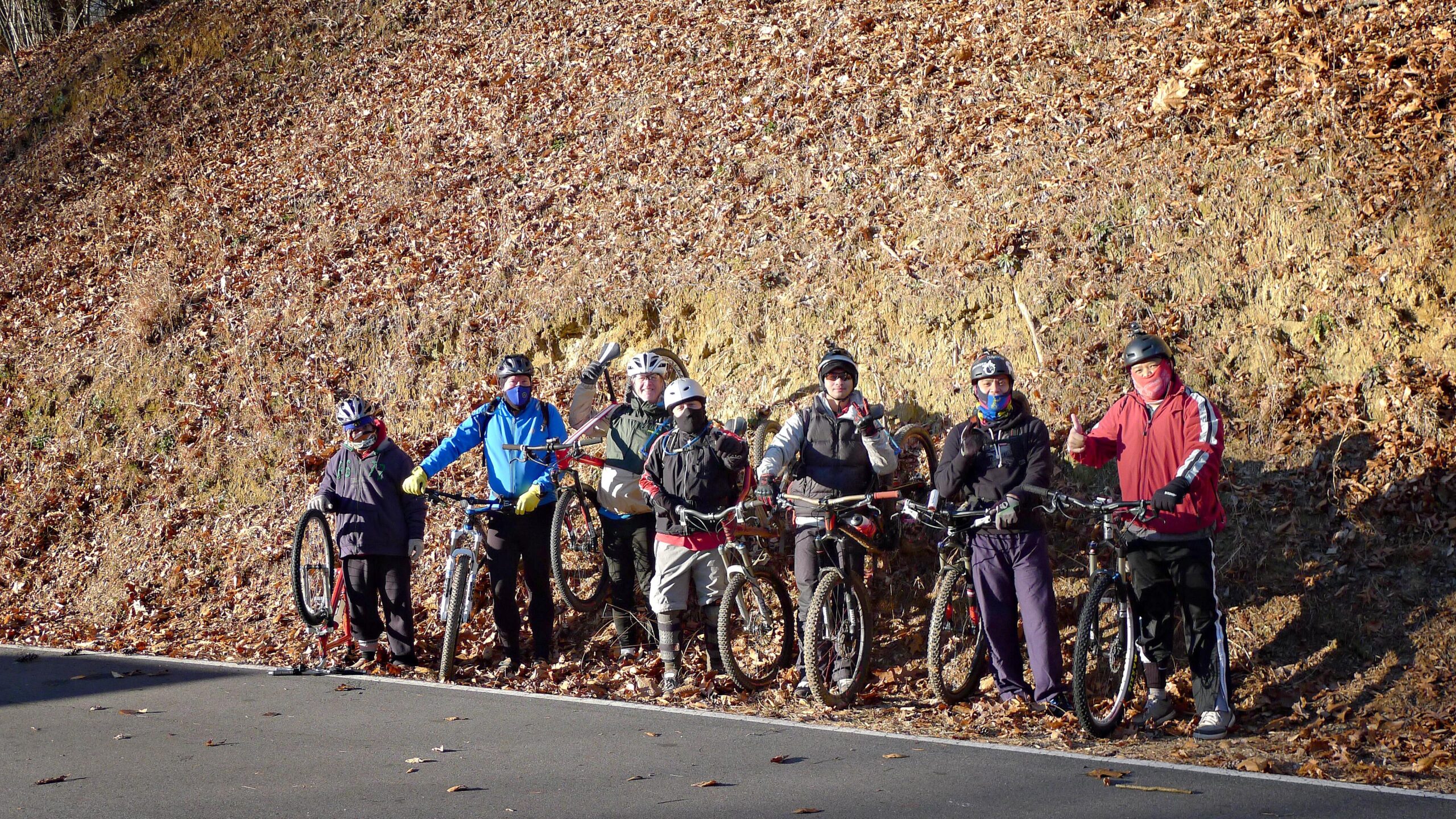 A group of seven mountain bikers standing together on a roadside, wearing helmets and biking gear. They are smiling and posing with their bicycles, surrounded by a hilly, leaf-covered landscape. The scene reflects a sunny day in autumn. Warrior Creek mountain bike trail.