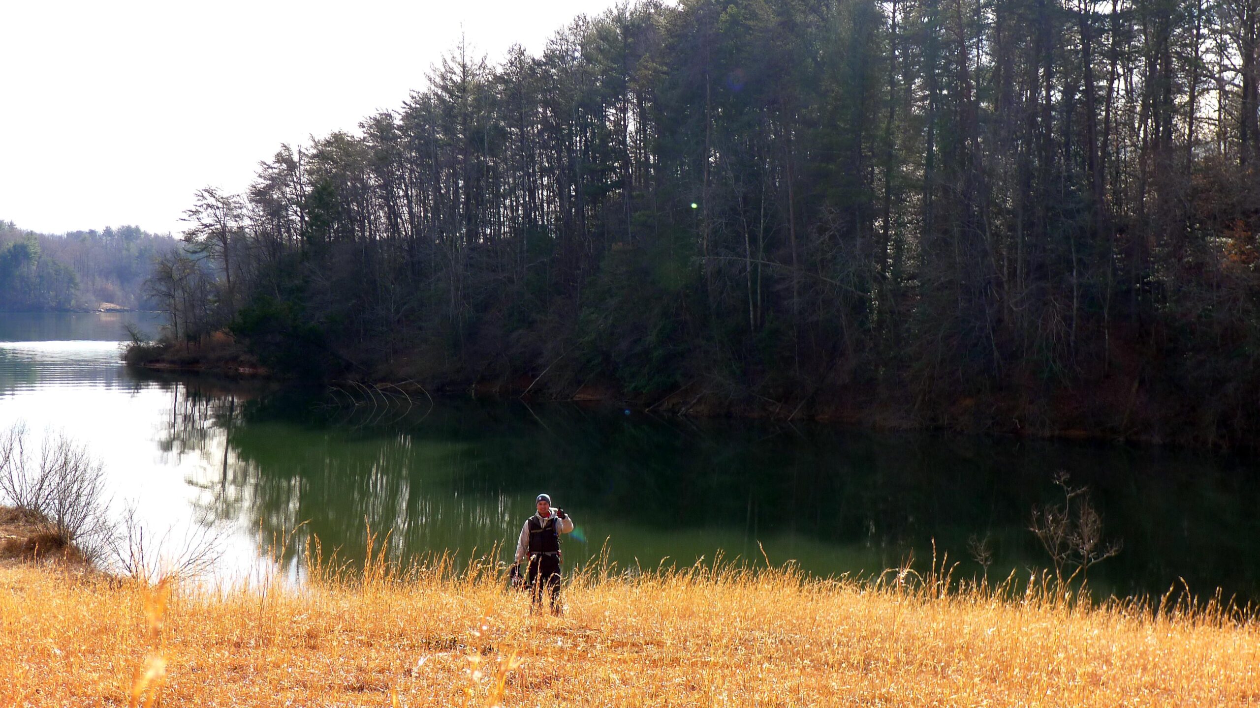 A person standing in a grassy area near a calm lake, surrounded by trees. The sunlight casts a soft glow, creating a serene atmosphere. The individual appears to be pausing or taking a moment to enjoy the view. Warrior Creek mountain bike trail.