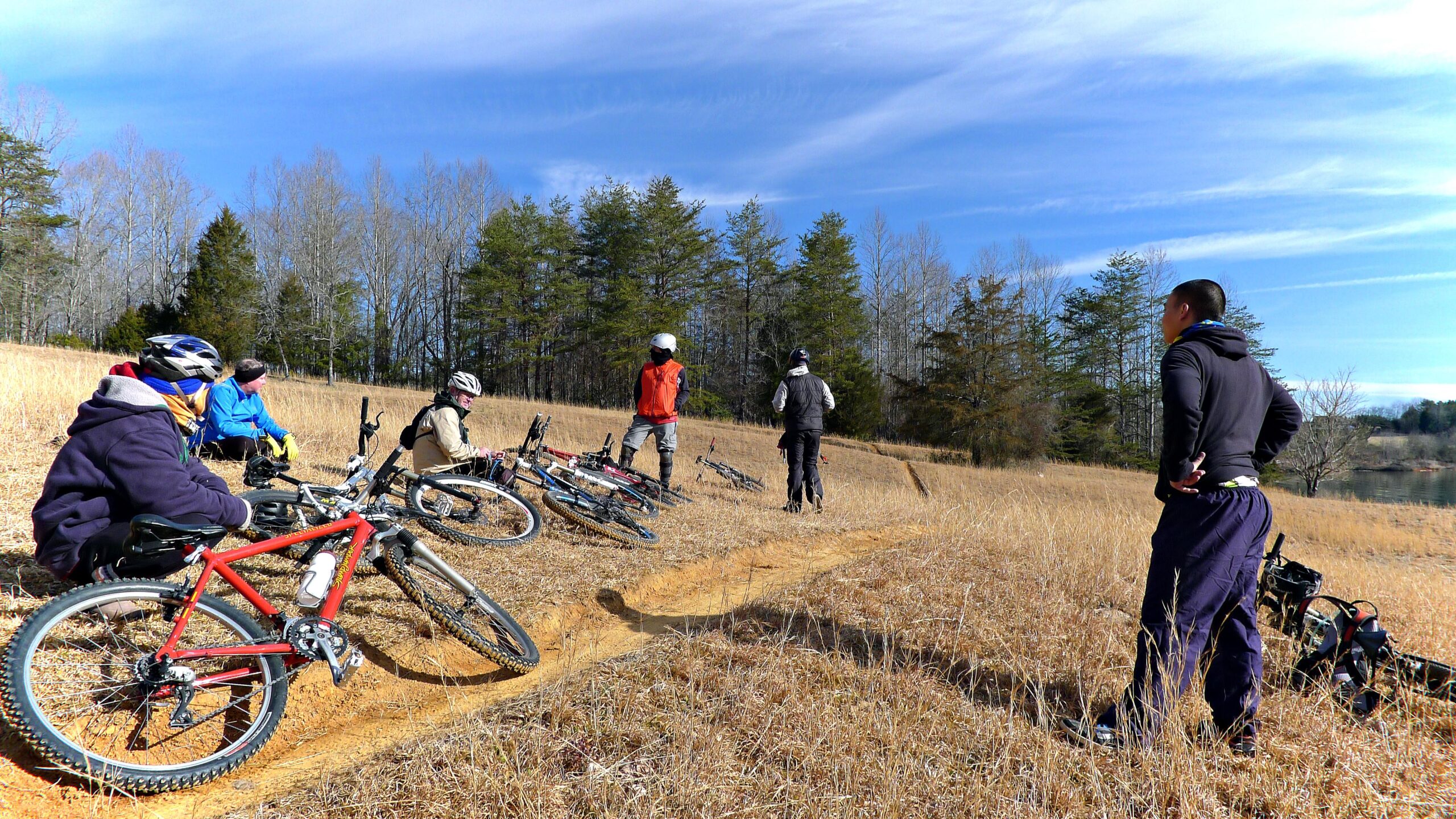 A group of five mountain bikers resting on a grassy hillside. Some are seated beside their bikes, while two are standing and looking out over the landscape. The scene is set against a backdrop of trees and a clear blue sky. Warrior Creek mountain bike trail.