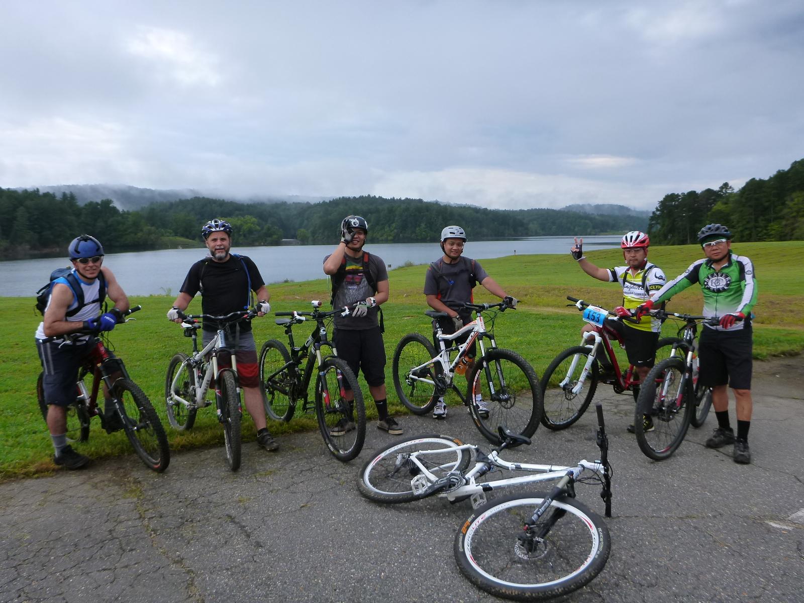 A group of six mountain bikers posing beside their bicycles near a lake, with a backdrop of green trees and cloudy skies. Two bicycles are lying on the ground, while the others are being held by the riders, who are smiling and making hand gestures to the camera. Dark Mountain Trail mountain bike trail.