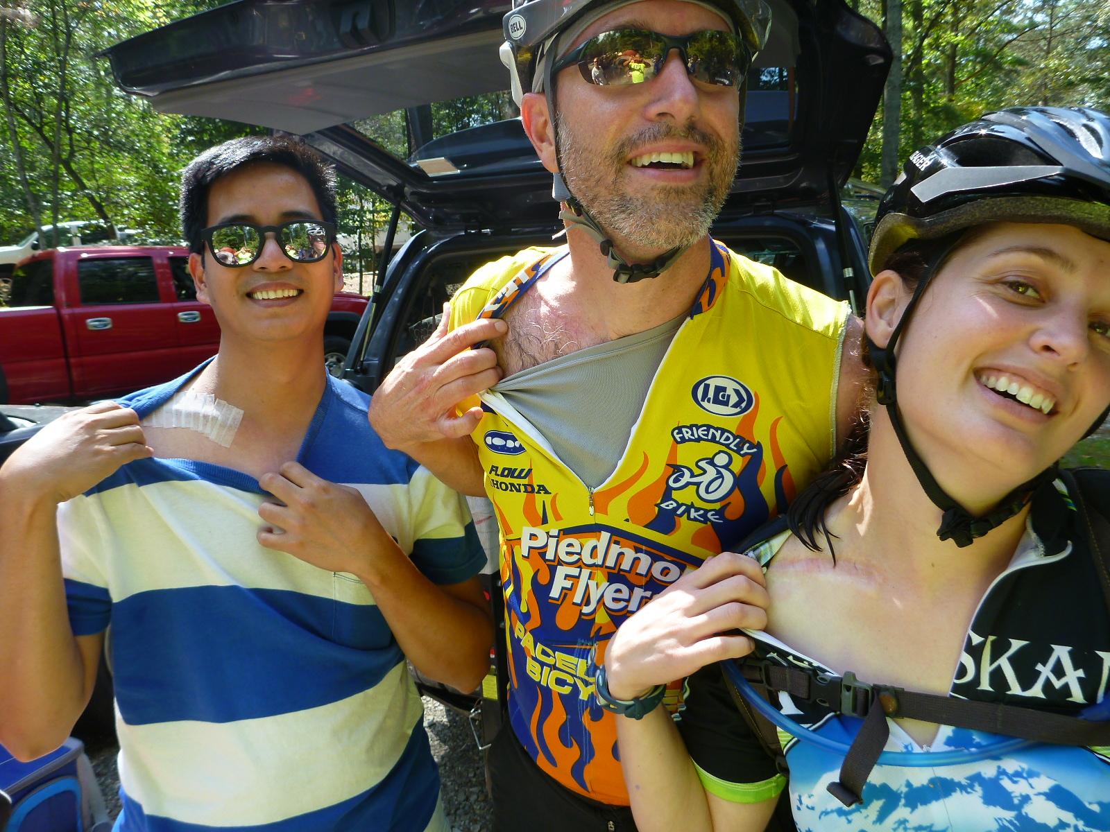 Three friends are displaying their upper bodies with smiles, showcasing their biking gear and small bandages on their shoulders. The background features trees and parked vehicles. The atmosphere appears light and cheerful, likely after a biking adventure. Warrior Creek mountain bike trail.