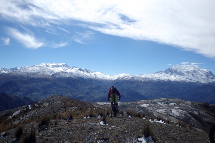 A person riding a mountain bike on a rocky ridge, overlooking a picturesque landscape of snow-capped mountains and valleys under a clear blue sky with wispy clouds.