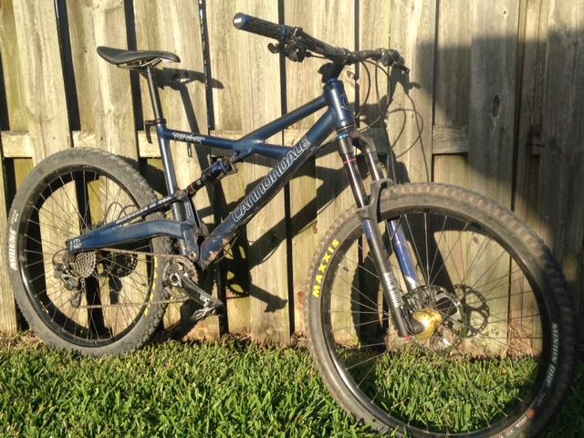 Cannondale Prophet: A blue Cannondale mountain bike leaning against a wooden fence, with visible knobby tires and a suspension system. The grass in the foreground is green and the bike is positioned in a way that showcases its frame and components.