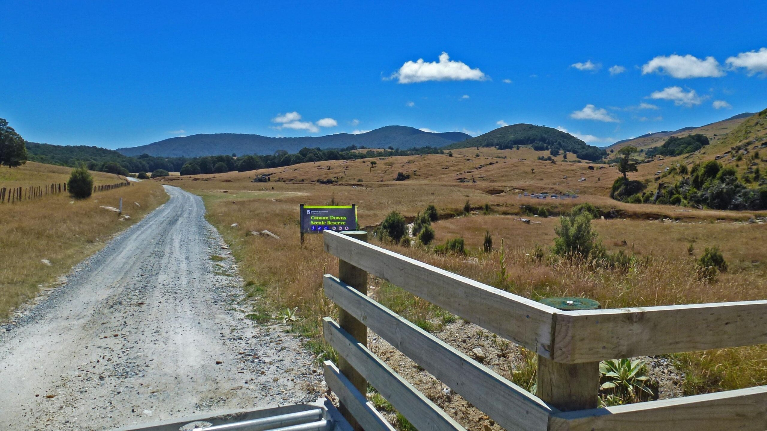 A gravel road leading into a scenic landscape filled with golden grass and rolling hills under a bright blue sky with a few fluffy clouds. A wooden fence marks the entrance to the Canaan Downs Scenic Reserve, with a sign visible that provides information about the area. Rameka Track mountain bike trail.