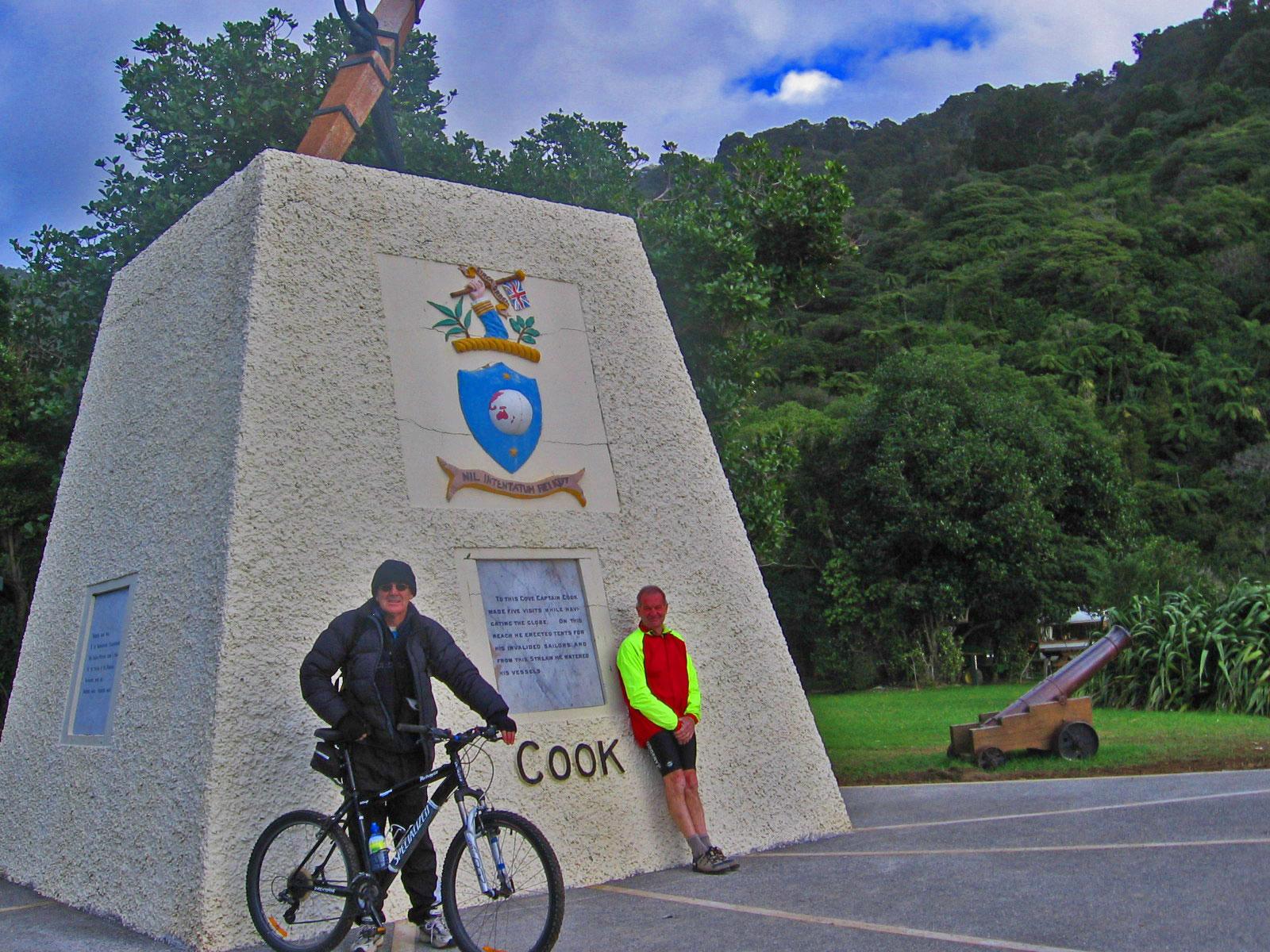 Two cyclists pose near a tall, white monument commemorating Captain James Cook. One rider stands beside a bicycle, wearing a black jacket and sunglasses, while the other leans against the monument in a bright red and yellow outfit. The background features lush greenery and a cloudy sky. Queen Charlotte Track mountain bike trail.