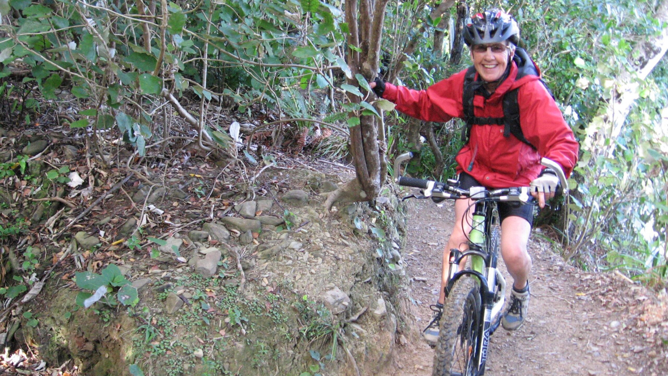 A person wearing a red jacket and helmet smiles while riding a mountain bike on a narrow dirt trail surrounded by greenery. Queen Charlotte Track mountain bike trail.
