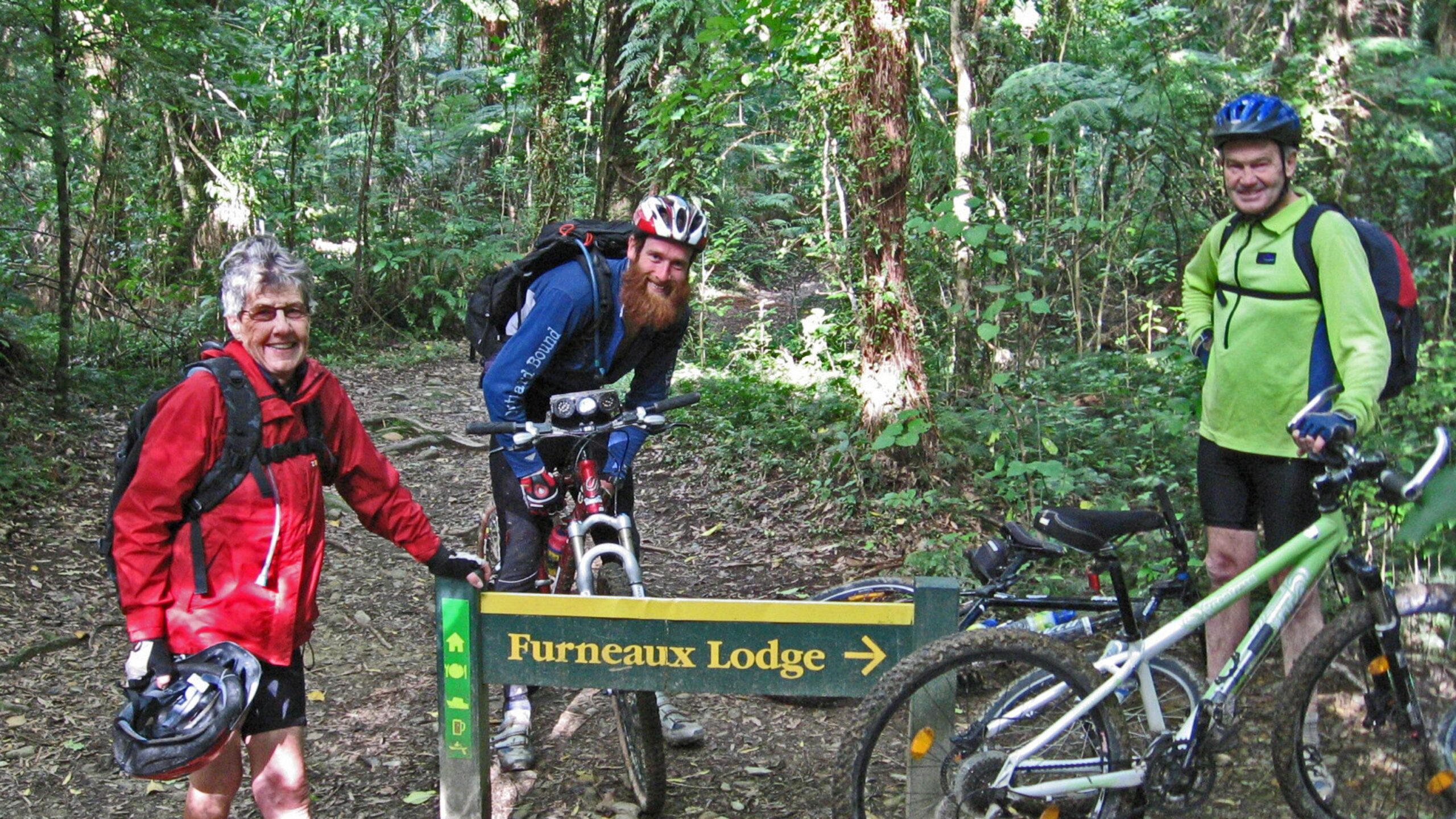 Three mountain bikers pause at a trail sign for Furneaux Lodge, surrounded by lush greenery in a forested area. One person in a red jacket smiles while holding a helmet, with a bike nearby. Another biker with a long beard sits on a bike, wearing a blue shirt, and the third biker wears a green jacket, standing next to his bike. The atmosphere is cheerful and outdoorsy. Queen Charlotte Track mountain bike trail.