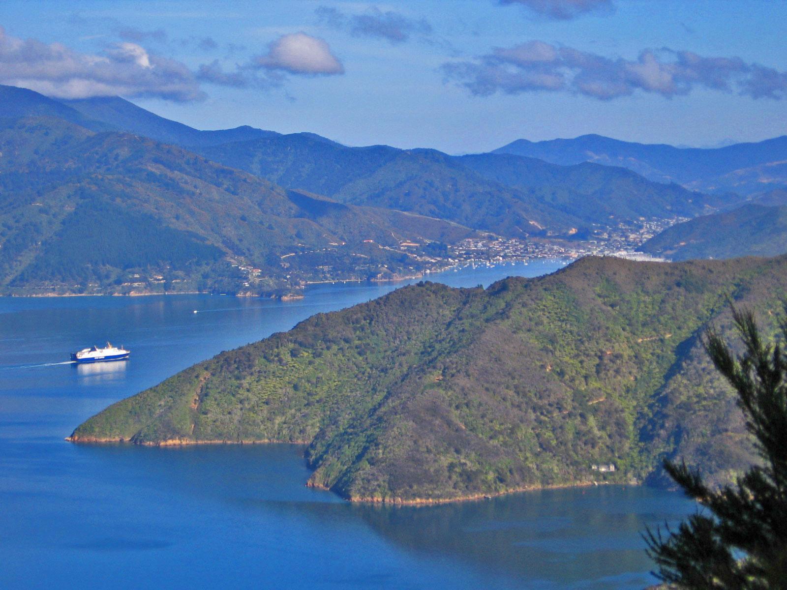 A scenic view of a coastal landscape featuring rolling hills and a blue bay. A ferry boat is seen navigating through the water, surrounded by lush greenery and distant mountains under a clear blue sky with a few scattered clouds. Queen Charlotte Track mountain bike trail.