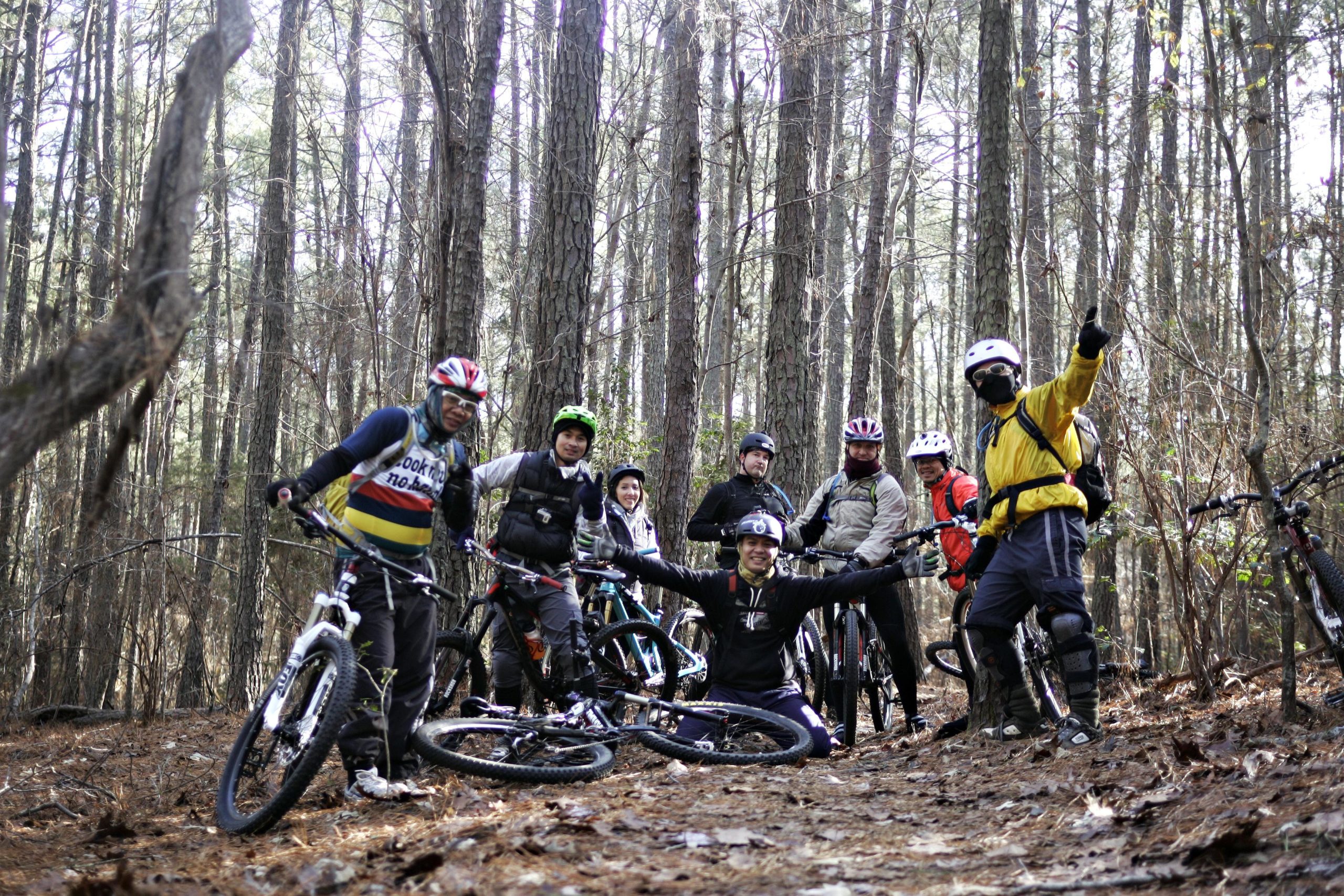 A group of eight mountain bikers posing together in a forest setting. The group is wearing helmets and biking gear while standing next to their bikes on a leaf-covered trail surrounded by tall trees. They appear cheerful and engaged, with some individuals giving thumbs up and others smiling toward the camera. Beaver Dam mountain bike trail.