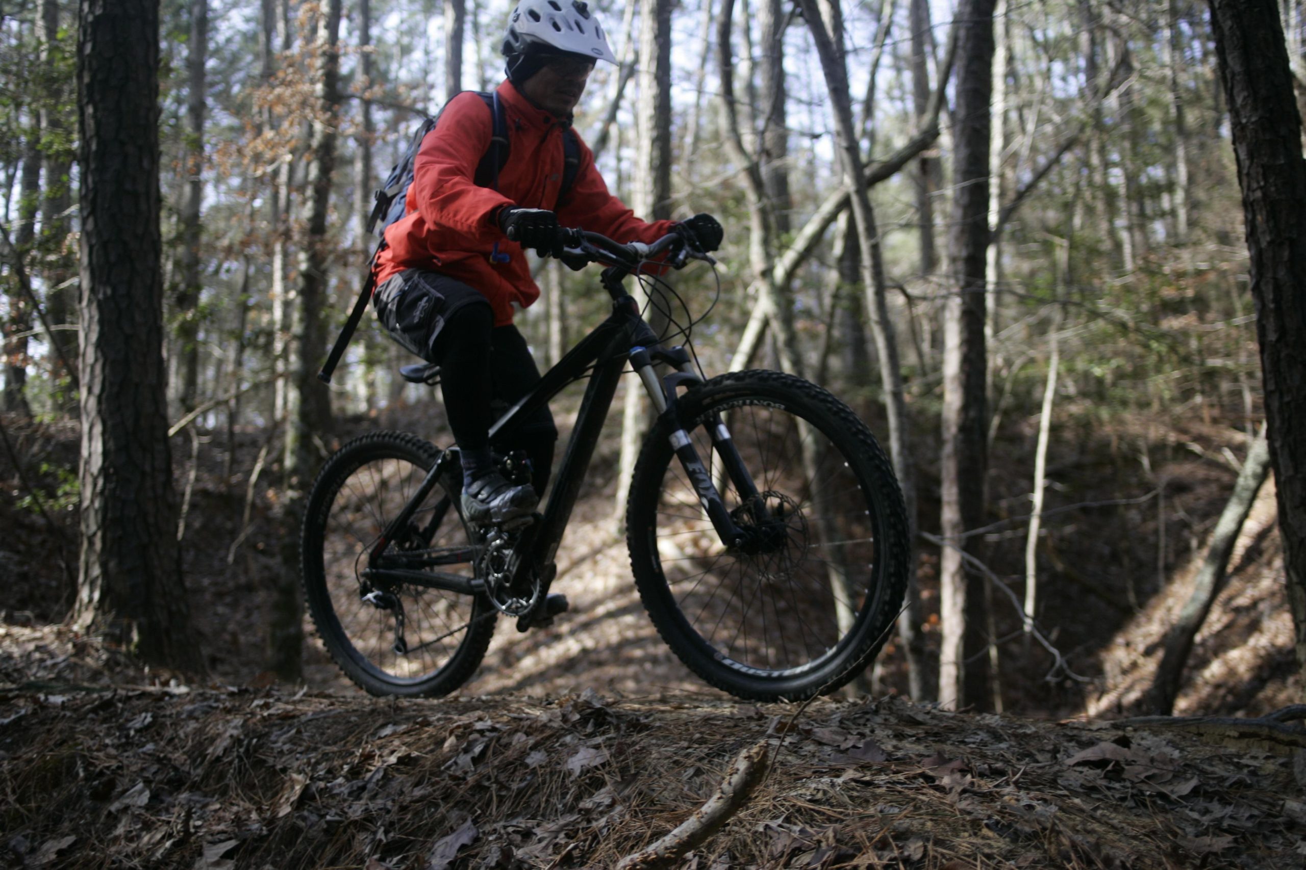 A mountain biker dressed in a bright orange jacket and black pants rides along a narrow, leaf-covered trail in a wooded area with tall trees in the background. The biker is focused, navigating a section of the trail with a slight incline and varied terrain. Beaver Dam mountain bike trail.