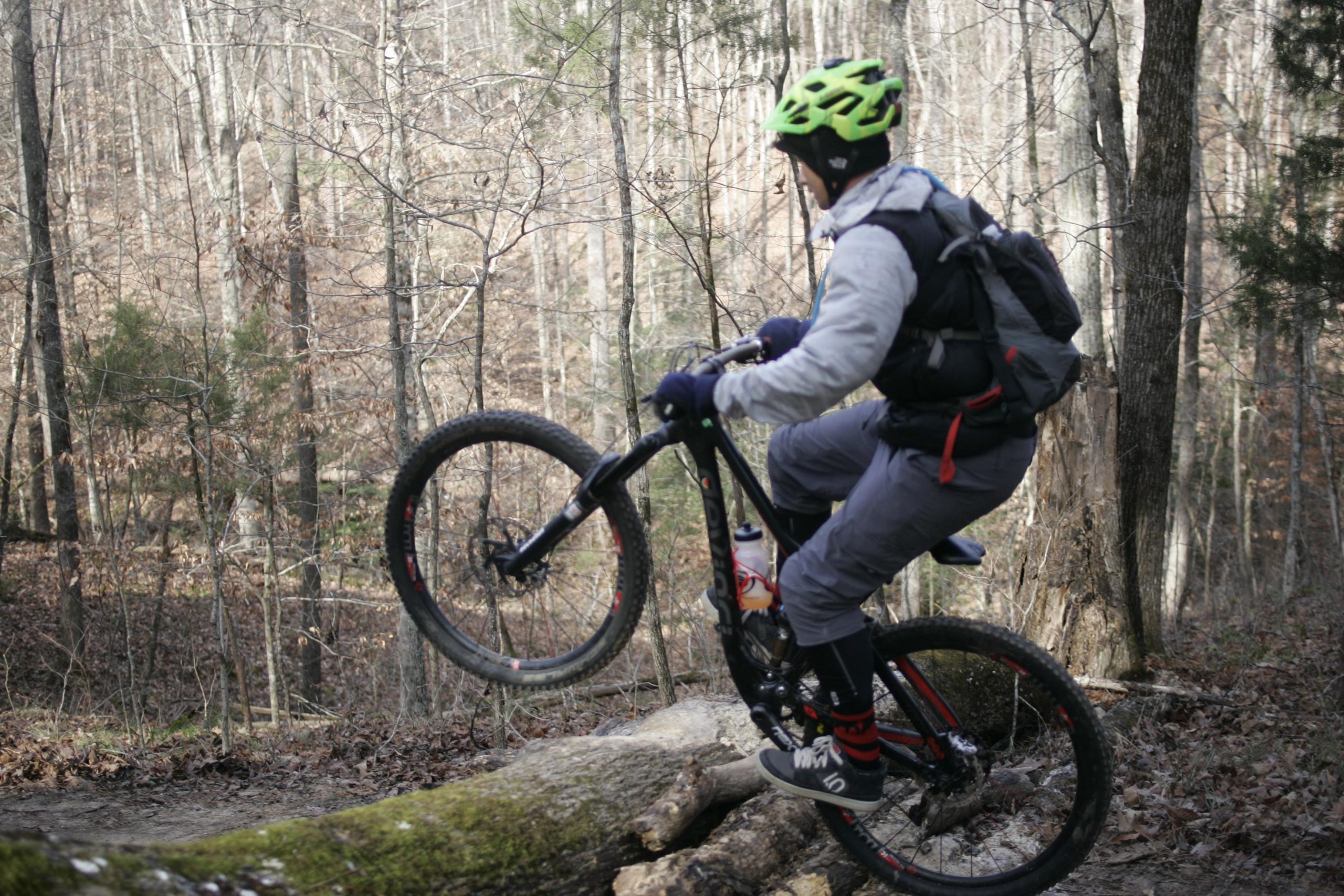 A person riding a mountain bike on a wooded trail, performing a wheelie over a fallen log. The rider is wearing a bright green helmet, gray outerwear, and black gloves, with a backpack and water bottle attached to the bike. The background features bare trees and a natural forest setting. Beaver Dam mountain bike trail.