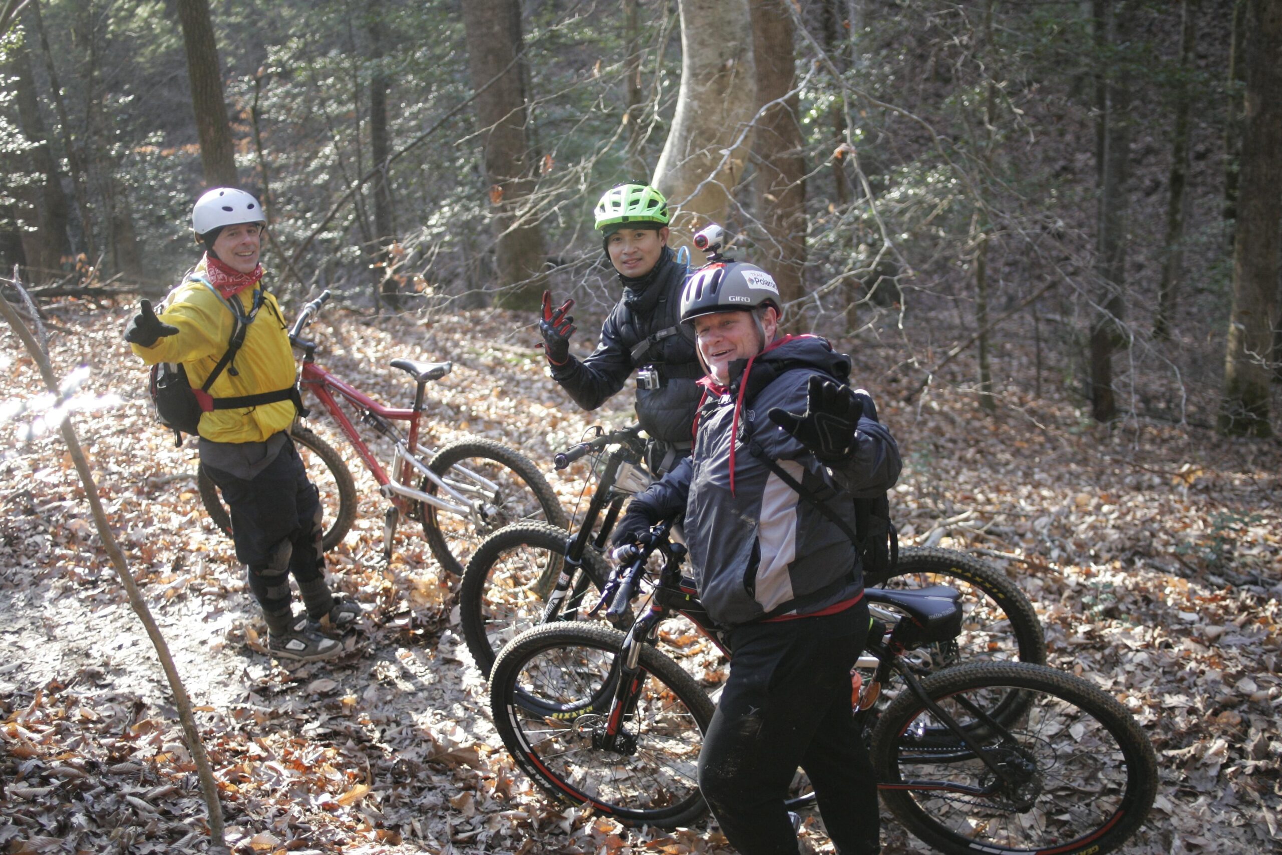 Three mountain bikers, wearing helmets and outdoor gear, pose for a photo on a leaf-covered trail in a wooded area. Two of them are holding up peace signs, while the third smiles and gives a thumbs-up. Nearby, their mountain bikes are parked on the ground amidst the fallen leaves and trees. The scene captures a lively moment in nature. Warrior Creek mountain bike trail.