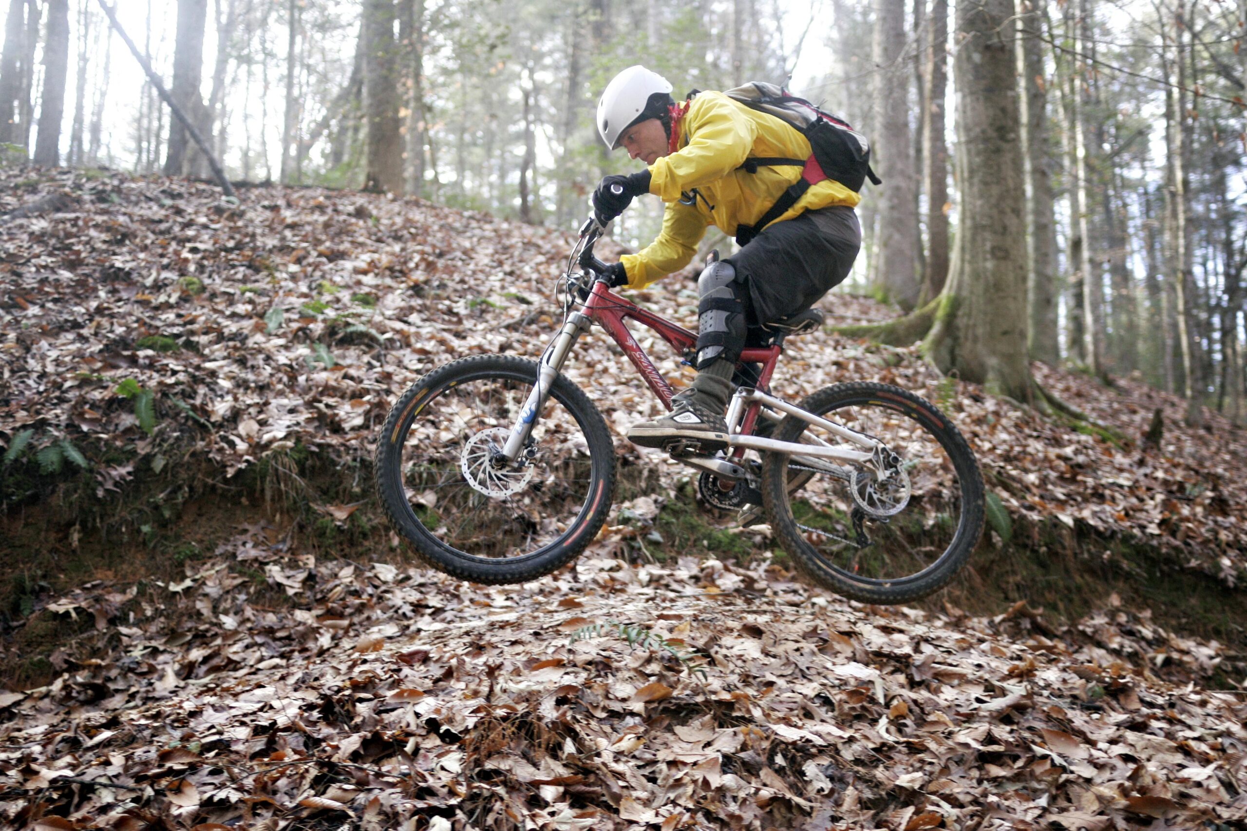 A mountain biker in a yellow jacket and helmet jumps over a small incline covered with fallen leaves in a wooded area. The rider is crouched down on a red mountain bike, showcasing an action shot in a natural outdoor setting. Warrior Creek mountain bike trail.