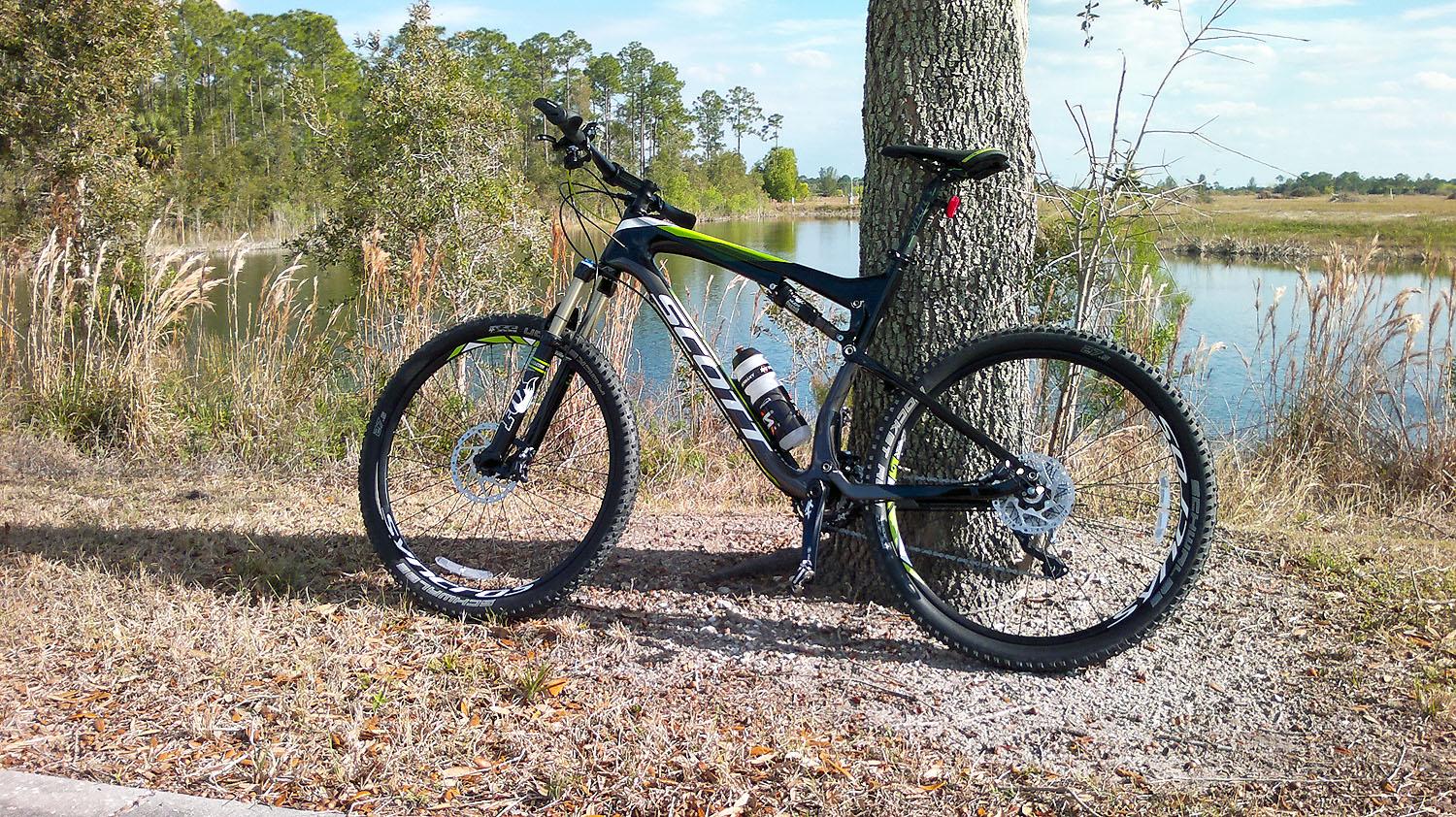 Scott Scott Spark 720: A black and green mountain bike leaning against a tree, with a calm body of water and trees in the background. The bike features thick tires and a water bottle mounted in its frame, surrounded by grass and small pebbles.