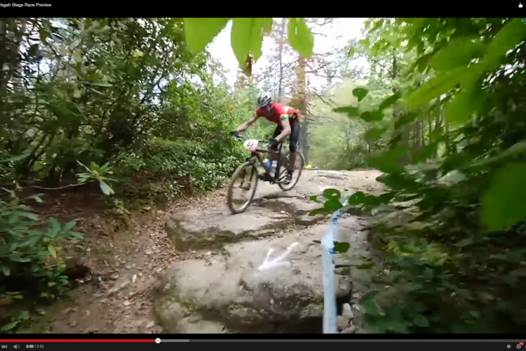 A mountain biker navigating a rocky trail surrounded by greenery, captured during the 2015 Pisgah Stage Race preview. The cyclist is in a dynamic riding position, with a race number visible on their bike.