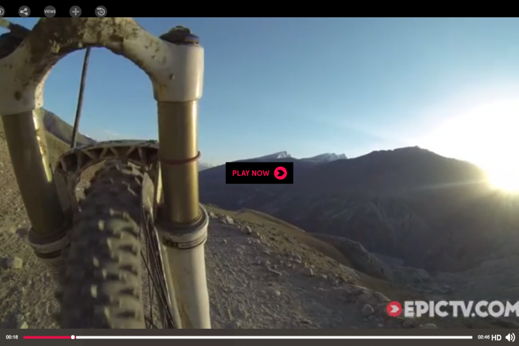 View from the front of a mountain bike, showcasing the handlebars and tire as it rides along a rocky path, with a scenic mountain landscape and sunset in the background. A "Play Now" button is visible on the screen.