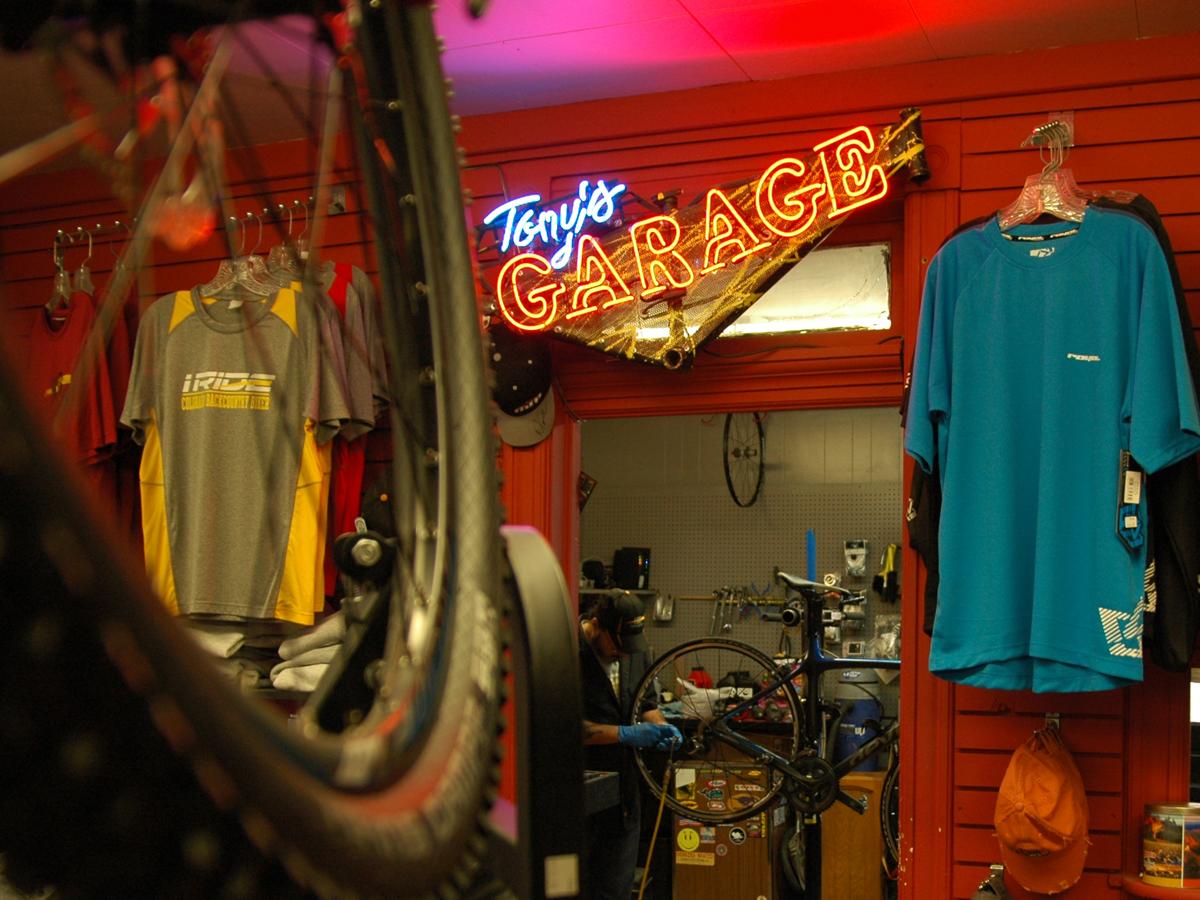 A cozy bike shop interior featuring a neon sign reading "Tony's GARAGE." In the foreground, a bicycle wheel is prominently displayed, while colorful cycling shirts hang on the walls. The background shows a workshop area with tools and bike parts.