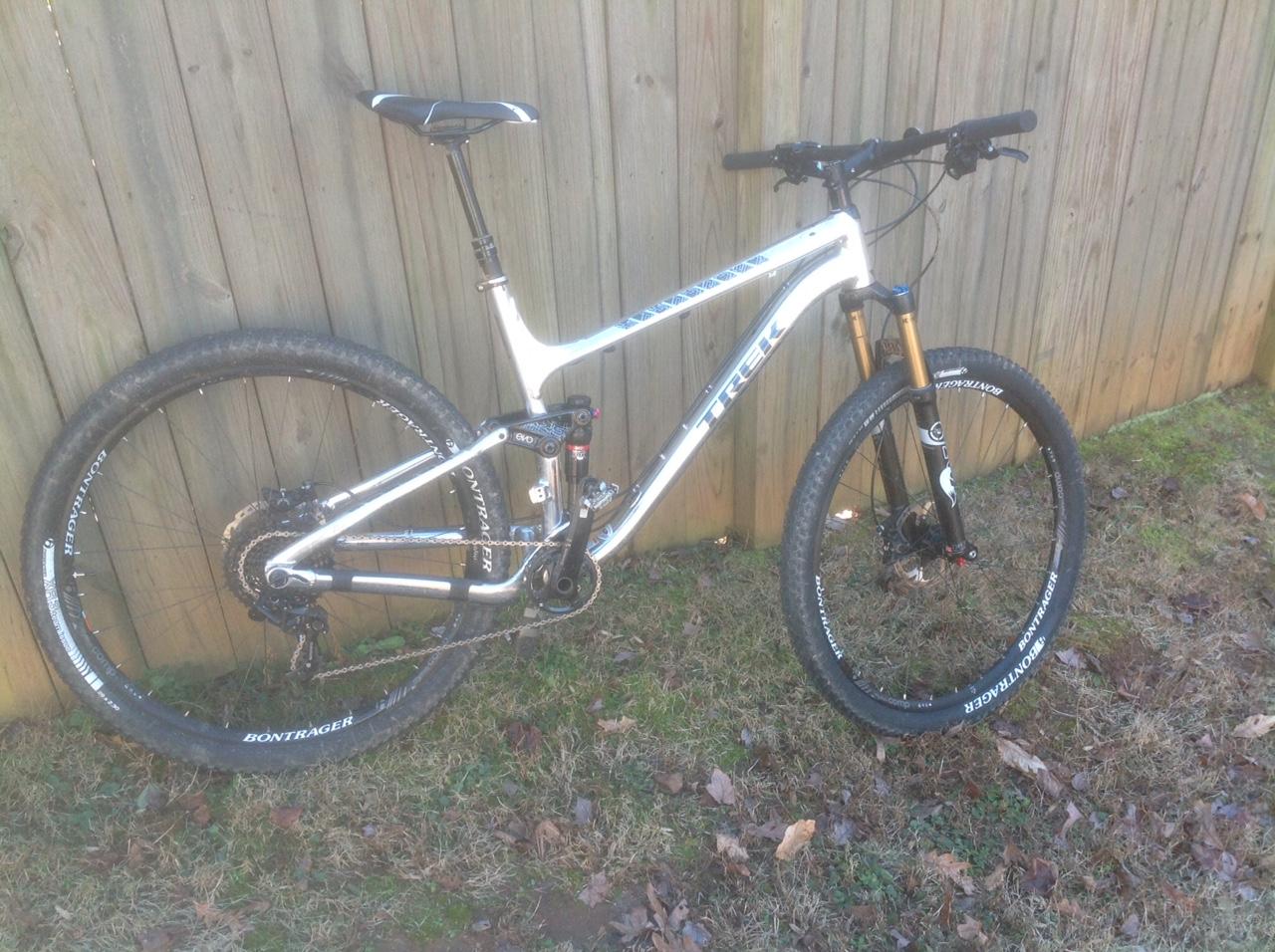 Trek Fuel EX 9 29 XO1: A silver and black mountain bike leaning against a wooden fence, featuring prominent wheels with Bontrager tires and a sleek aluminum frame. The bike has a suspension fork and modern components, set on a grassy area with scattered leaves.