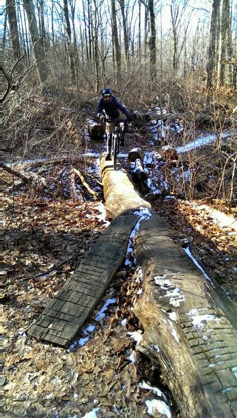 A cyclist navigating across a fallen tree trunk in a wooded area, surrounded by bare trees and scattered leaves. The ground is partially covered with snow, indicating a cold season. The cyclist is dressed in winter gear, focused on balancing while riding on the log. White Clay Creek mountain bike trail.