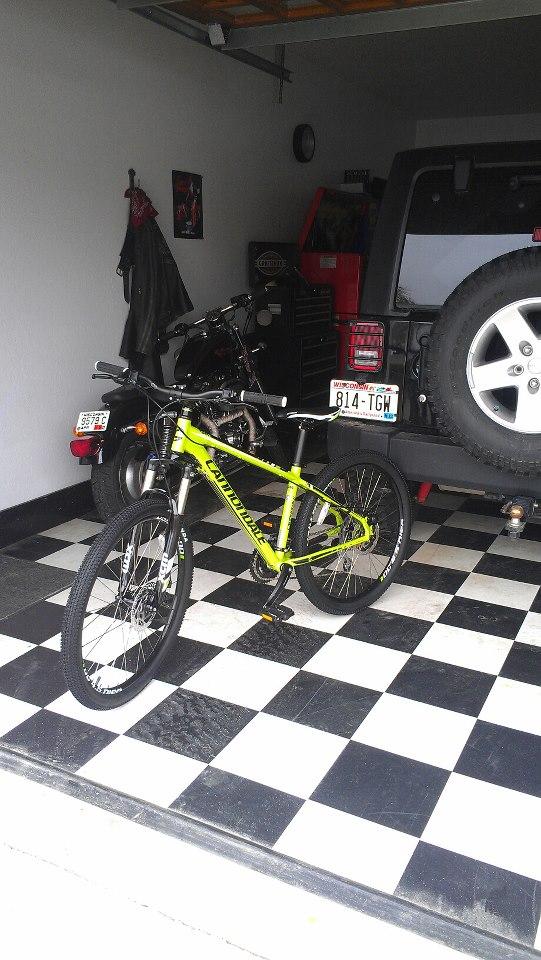 Cannondale Trail 5: A bright yellow mountain bike stands on a black and white checkered floor inside a garage. A motorcycle is parked in the background, alongside a toolbox. The garage features white walls and a clock on the wall. A large tire from a vehicle is visible in the foreground, adding to the garage setting.