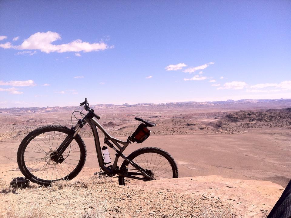 Specialized Camber Comp 29: A mountain bike leaning against a rock, overlooking a vast desert landscape with distant hills and a clear blue sky filled with a few clouds.