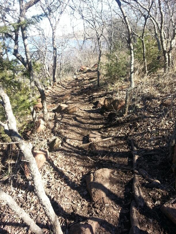 A narrow, winding trail through a wooded area, lined with rocks and trees. The path is covered in dry leaves, with branches overhead creating a natural archway. In the distance, a glimpse of water can be seen, suggesting proximity to a lake or river. The scene is bathed in natural light, indicating a clear day. Lake Lawtonka Trails mountain bike trail.