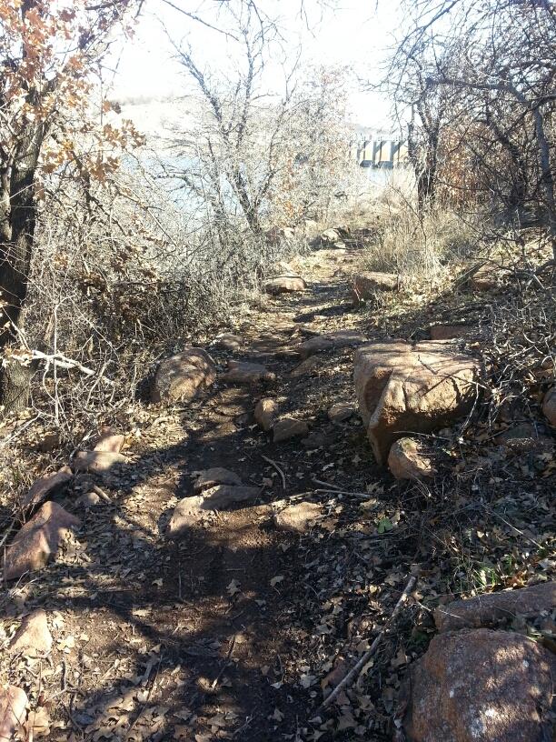 A narrow dirt path edged by rocks and overgrown vegetation, leading alongside a body of water. The scene features scattered dry leaves and bare branches, suggesting a late autumn setting. The horizon shows a faint outline of a dam or bridge structure in the distance. Lake Lawtonka Trails mountain bike trail.