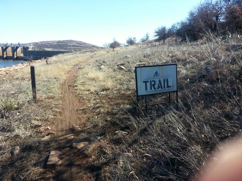 A dirt path leading through tall grass with a sign that says "TRAIL" on the right side. In the background, a body of water and a dam structure can be seen, framed by rolling hills and sparse trees under a clear blue sky. Lake Lawtonka Trails mountain bike trail.