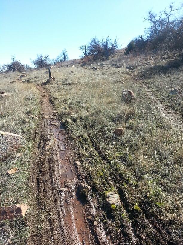 A dirt trail winding through a grassland area, marked by muddy tire tracks and scattered rocks. The scene is set against a clear blue sky, with sparse trees visible in the background. Lake Lawtonka Trails mountain bike trail.