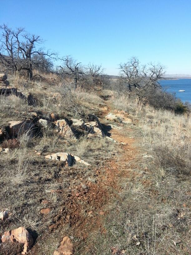 A narrow dirt path winding through rocky terrain, surrounded by sparse trees and grass. A clear blue sky is visible in the background, and a glimpse of water can be seen to the right, suggesting a nearby lake or river. Lake Lawtonka Trails mountain bike trail.
