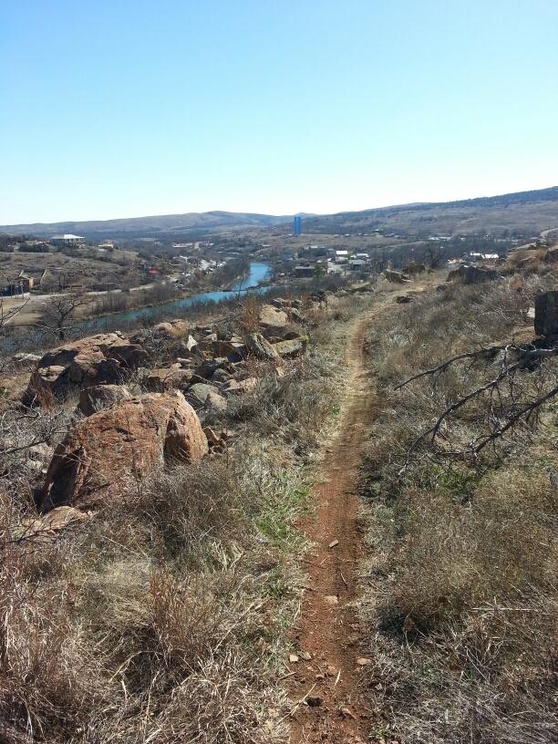 A winding dirt trail leading through rocky terrain and dry grass, overlooking a river and a small town in the distance under a clear blue sky. Lake Lawtonka Trails mountain bike trail.