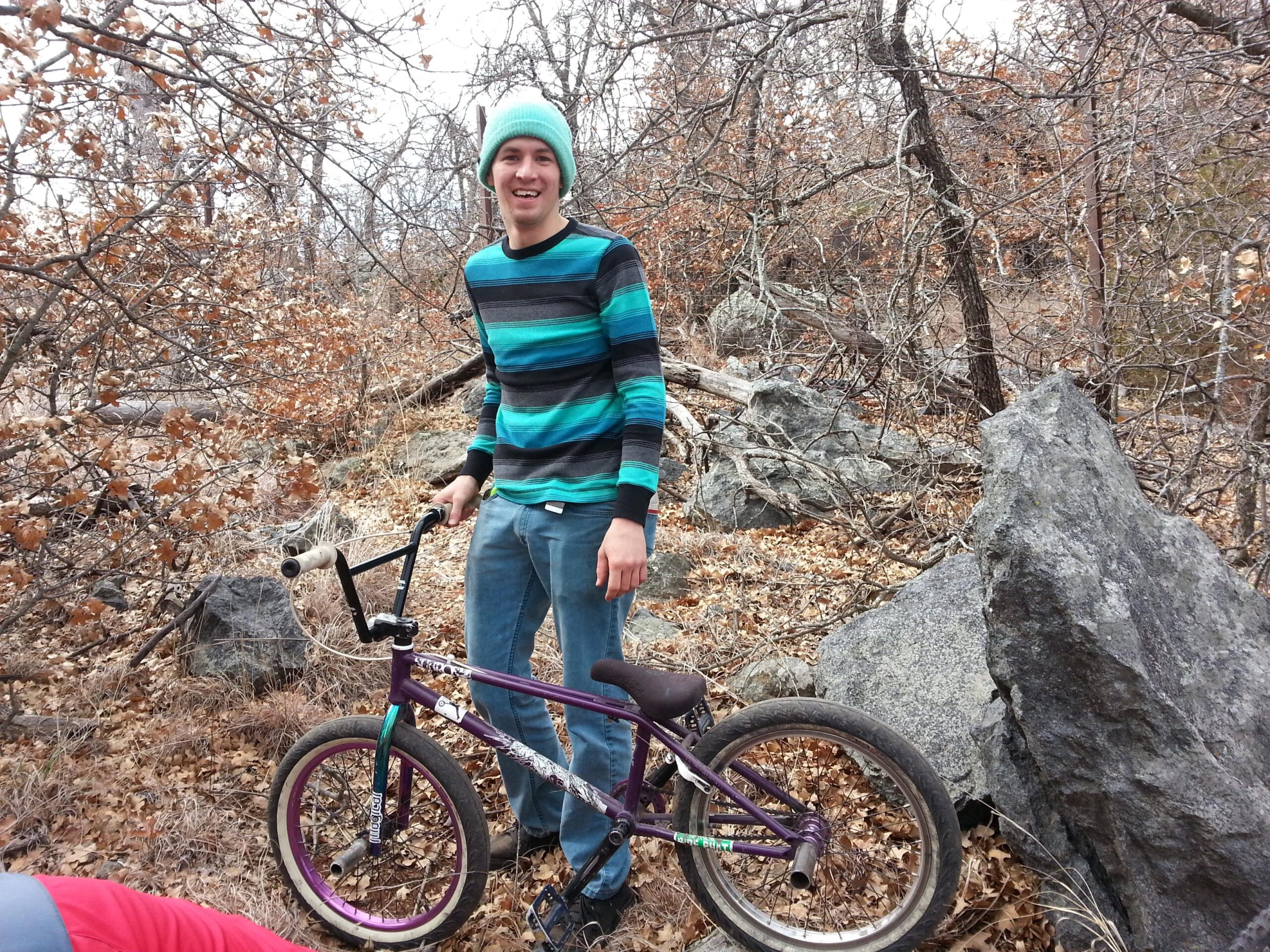 A young man wearing a teal and black striped sweater and a light blue beanie stands next to a purple BMX bike in a wooded area with rocks and dry leaves. The background features bare trees and rocky terrain, suggesting a natural outdoor setting. The man is smiling and appears cheerful, holding the bike's handlebars. Lake Lawtonka Trails mountain bike trail.