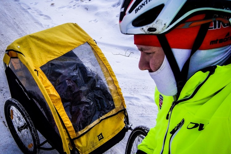 A cyclist wearing a bright yellow jacket and a helmet is pulling a child trailer with a transparent cover through a snowy landscape. The path is covered in white snow, and the cyclist is focused on the journey ahead.