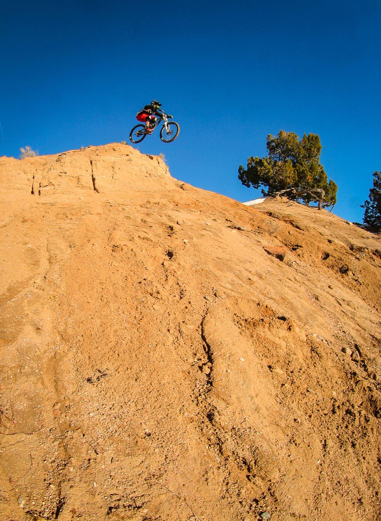 A mountain biker in mid-air is jumping off a steep sandy slope against a clear blue sky. A tree is visible in the background.